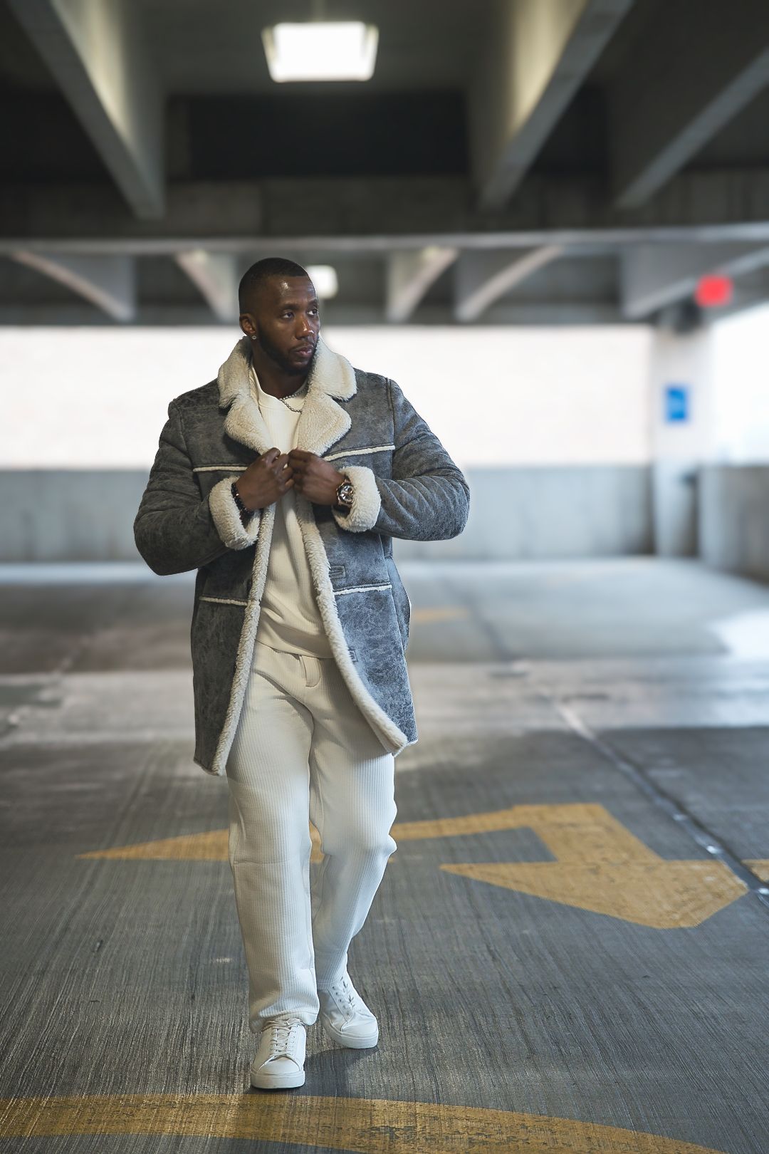 Person walking through parking garage wearing cream outfit and gray shearling coat.