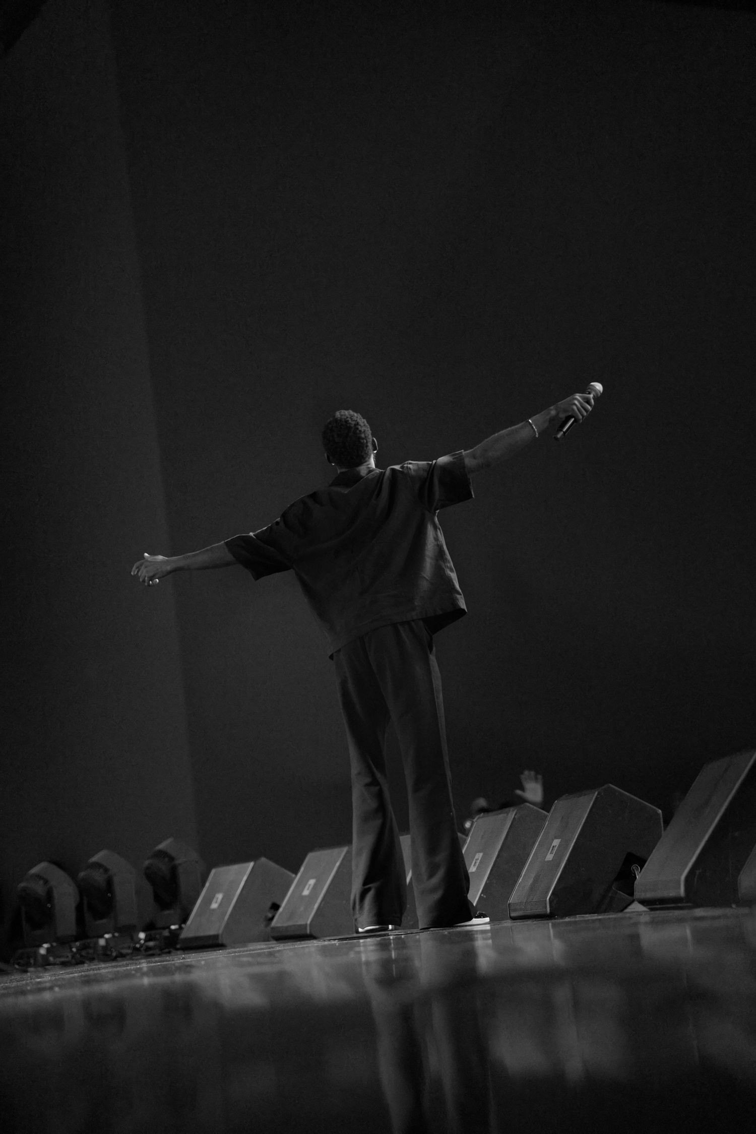 A silhouetted figure stands with arms outstretched on a dimly lit stage with chairs in the background.