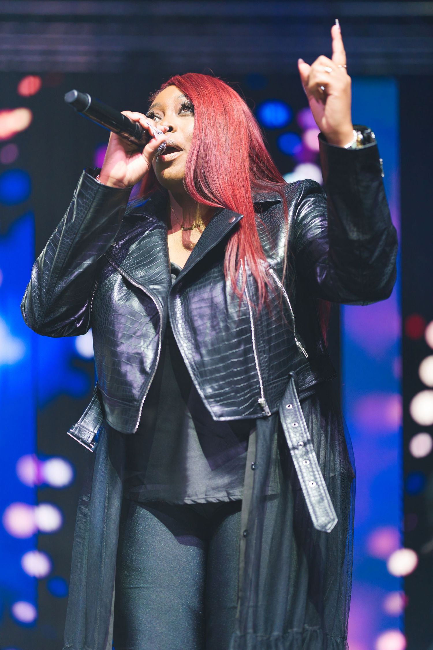 Performer with pink hair in leather jacket singing on stage with blue lighting effects.