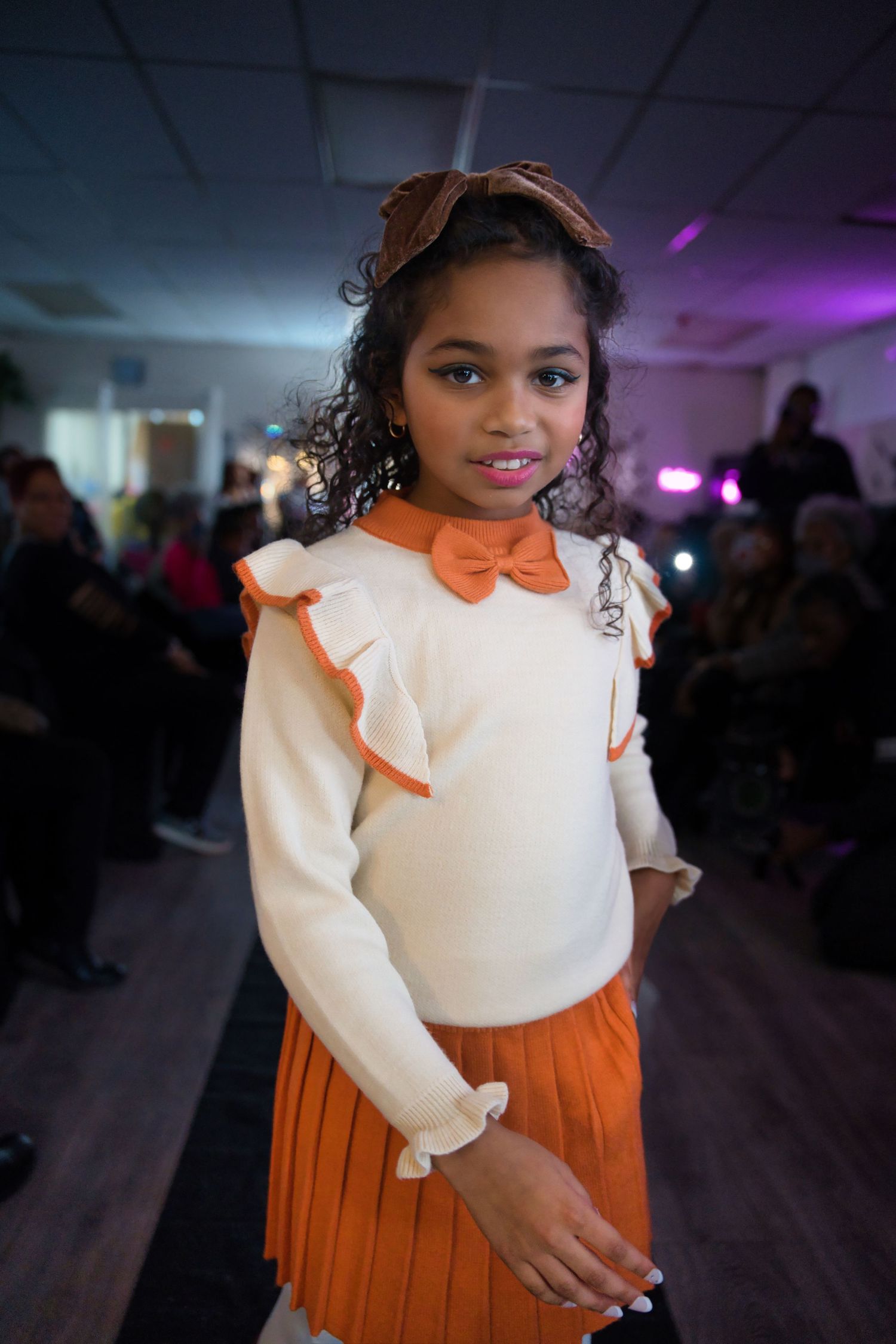 Young model wearing white top with orange bow and matching pleated skirt at fashion event.