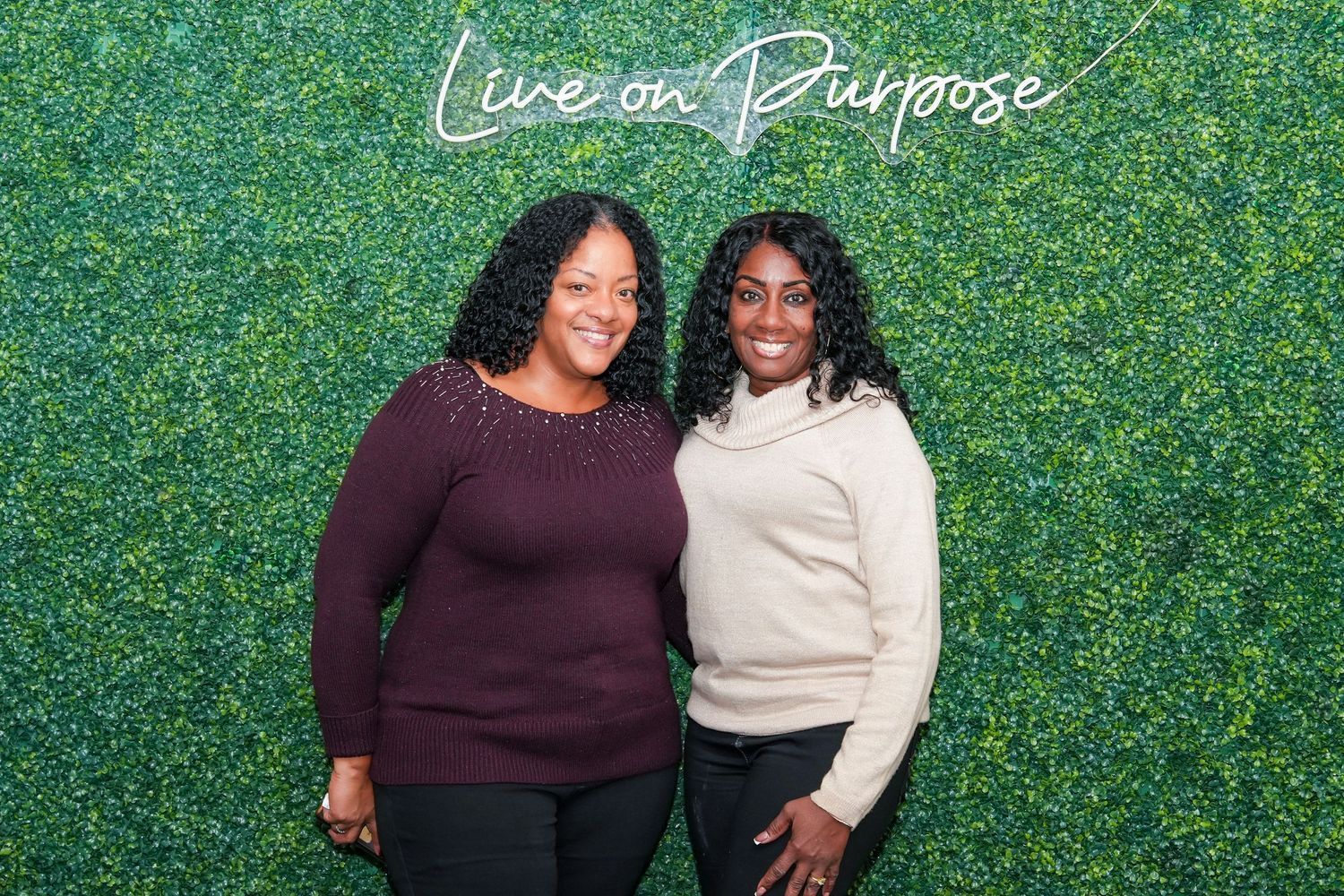 Two women in coordinating sweaters share a joyful moment against a green hedge backdrop.