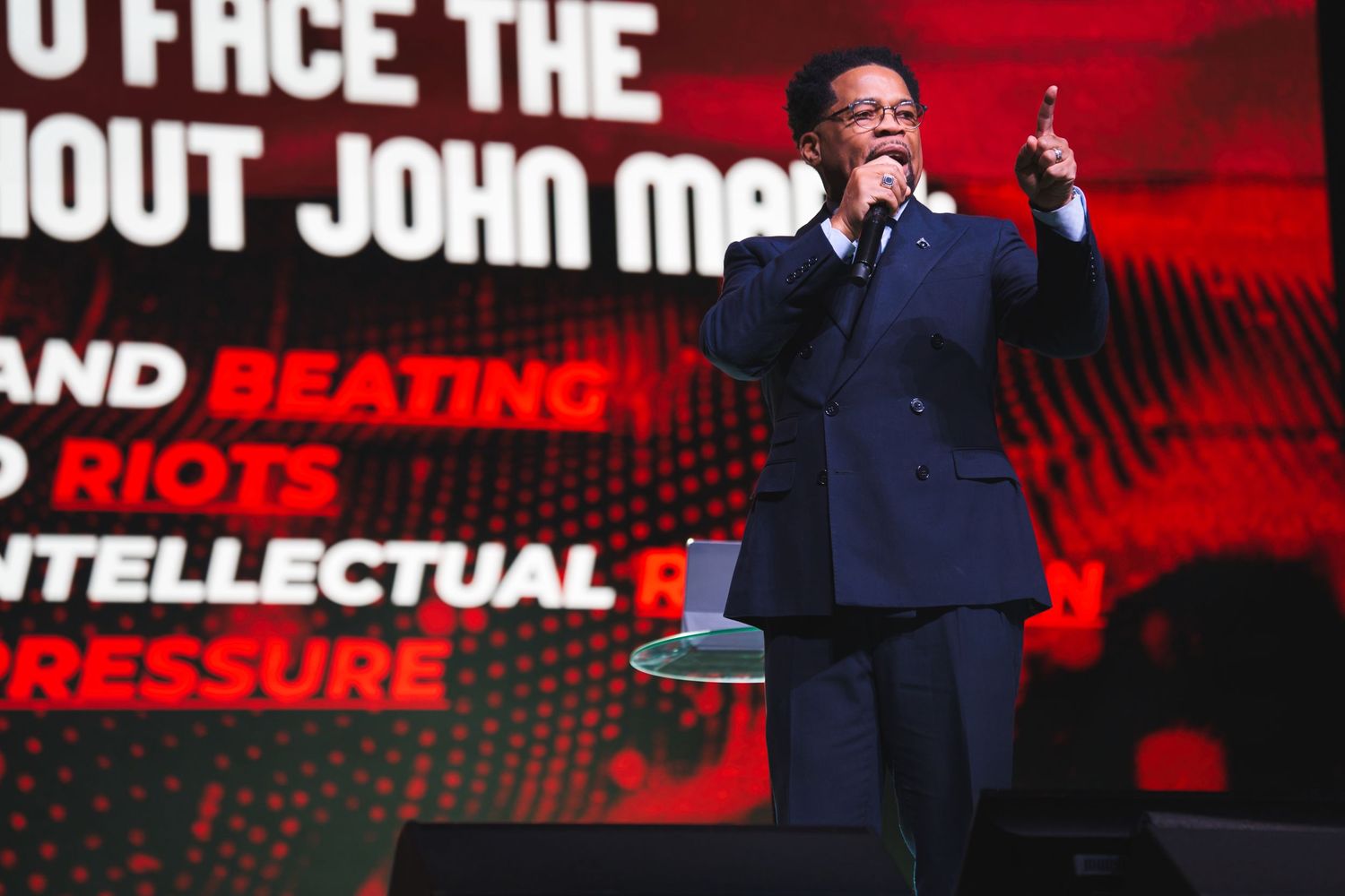 Presenter speaking at event with red digital backdrop and text overlays.