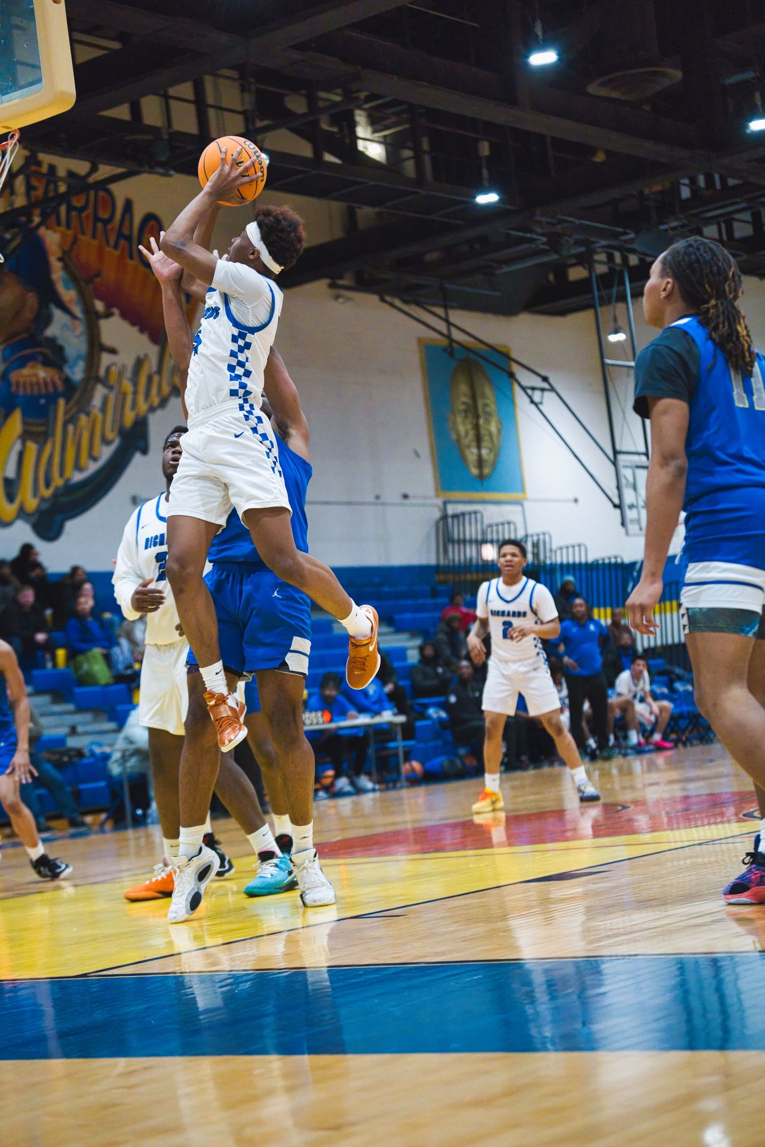 Basketball player leaps high to make a shot during an intense indoor game with blue court and spectators watching.