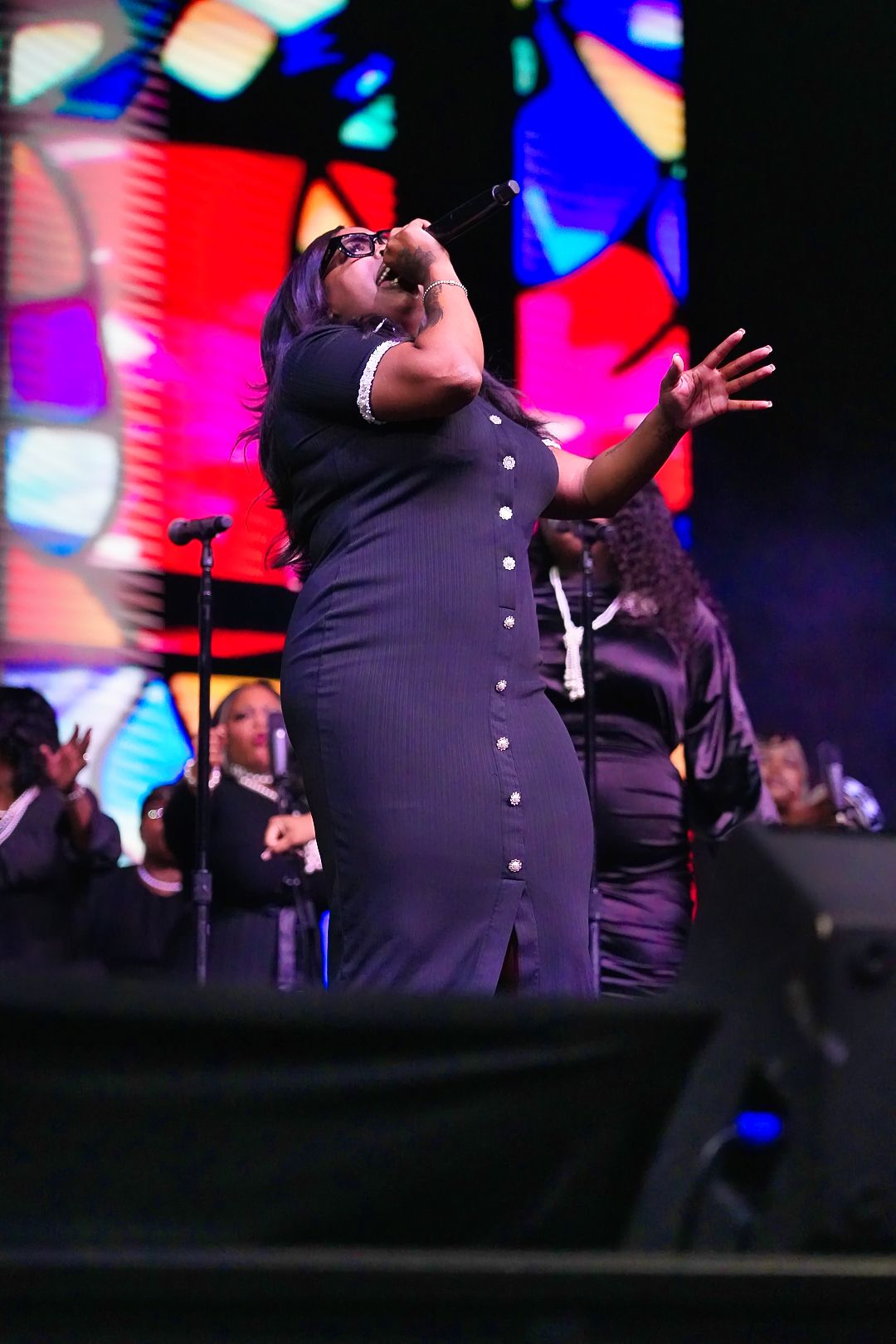 A performer in a black dress sings passionately on stage with colorful backdrop lighting.