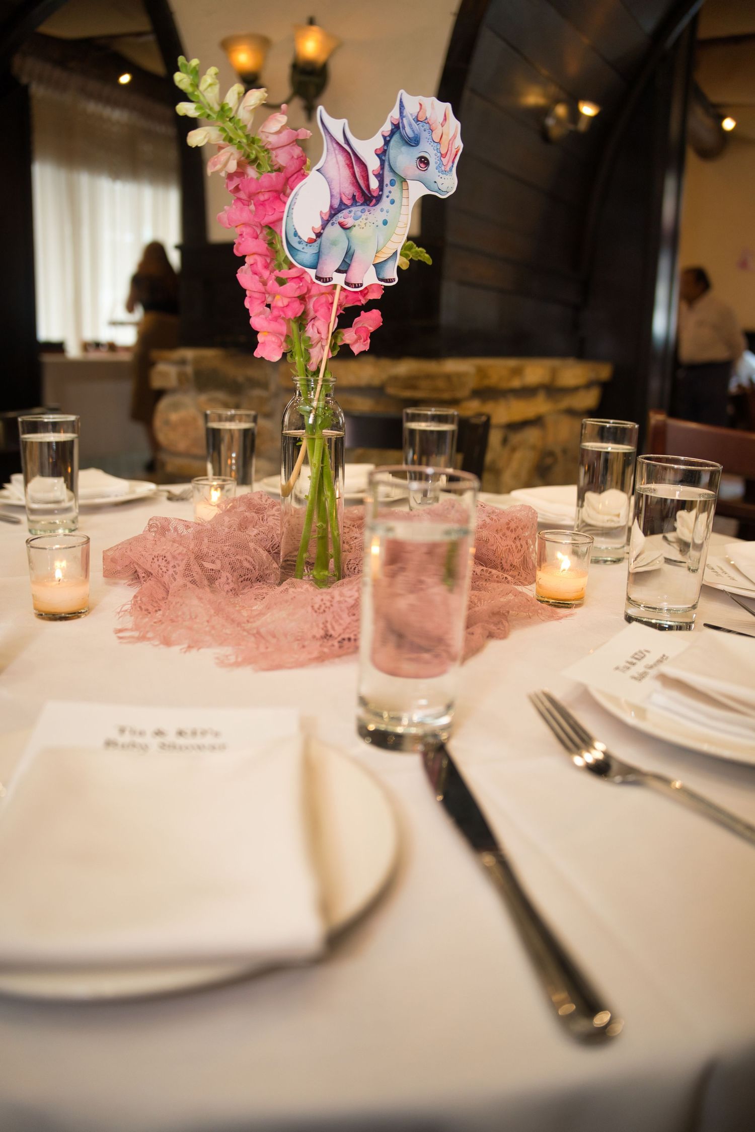 Wedding reception table setting with pink floral centerpiece, candles, and white plates on elegant tablecloth.