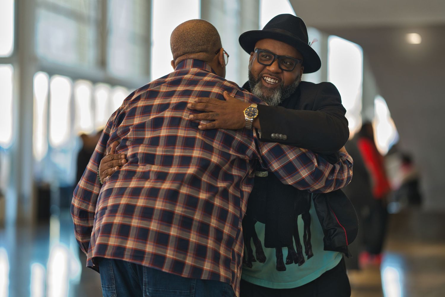 Two men embrace in a friendly hug in a hallway, one wearing a plaid shirt and the other in a black hat and sweater.