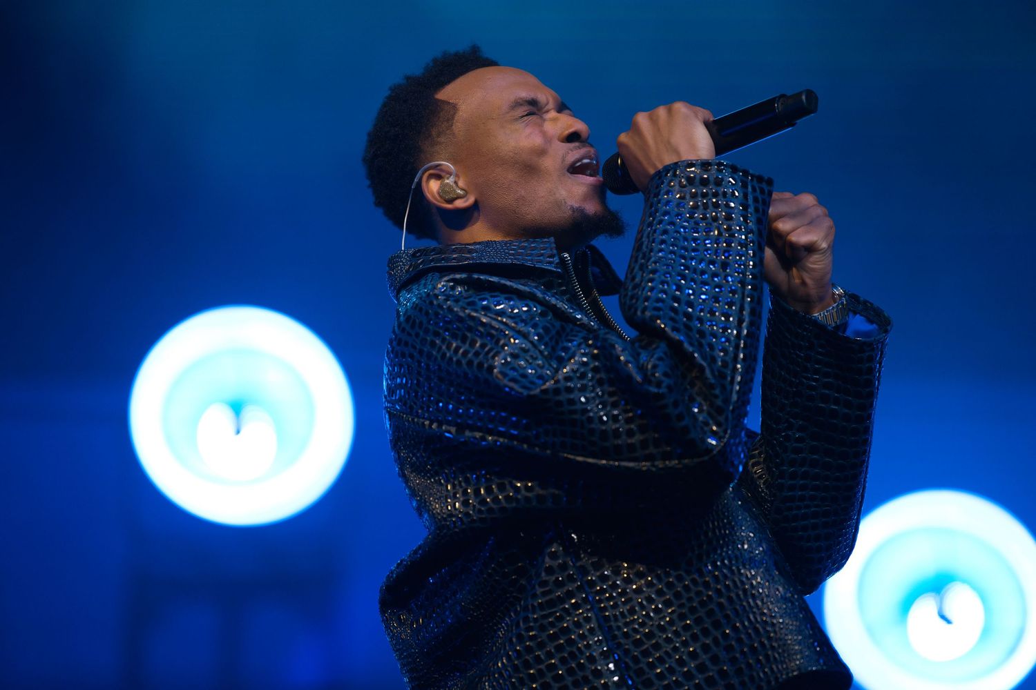 A singer performs energetically in a black and sparkly outfit against a blue-lit stage backdrop.