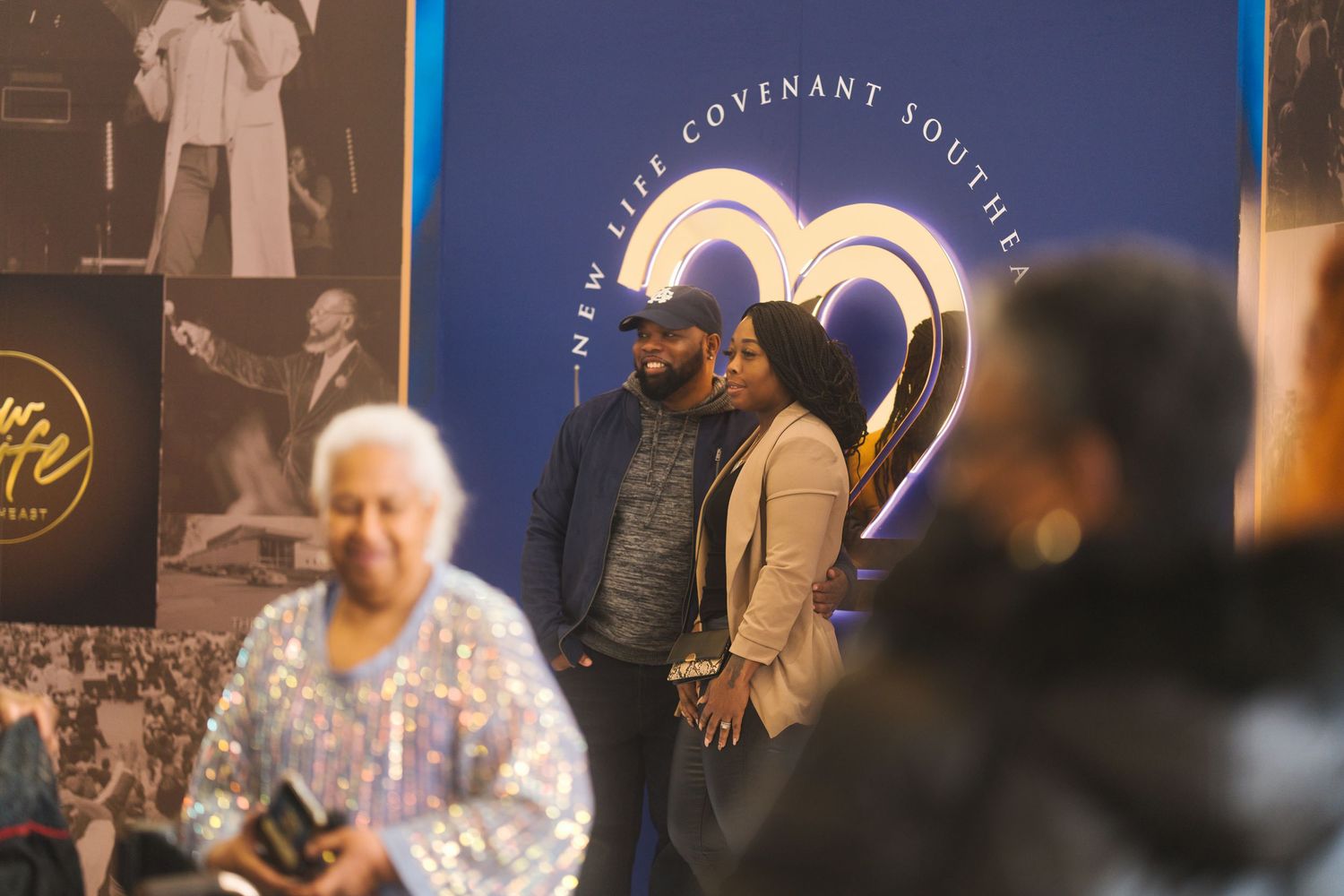 People gather at an event with a blue illuminated wall displaying a curved design in the background.
