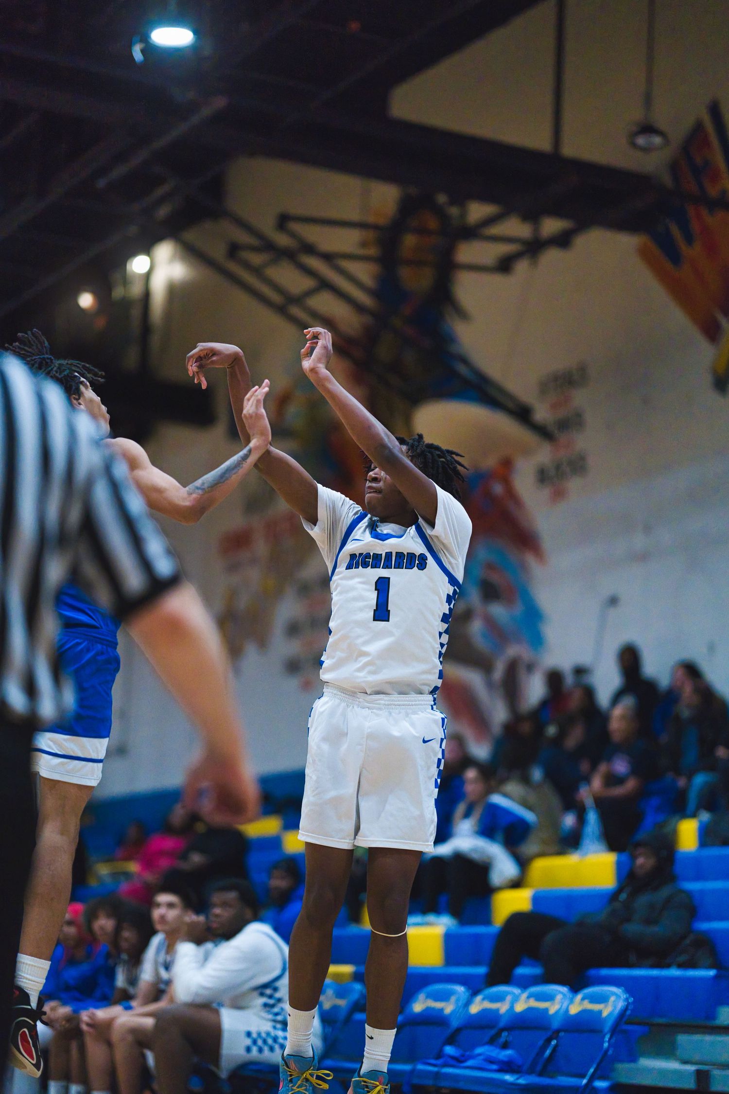 Basketball players in white and blue uniforms compete during an intense game in a gymnasium with blue stadium seating.