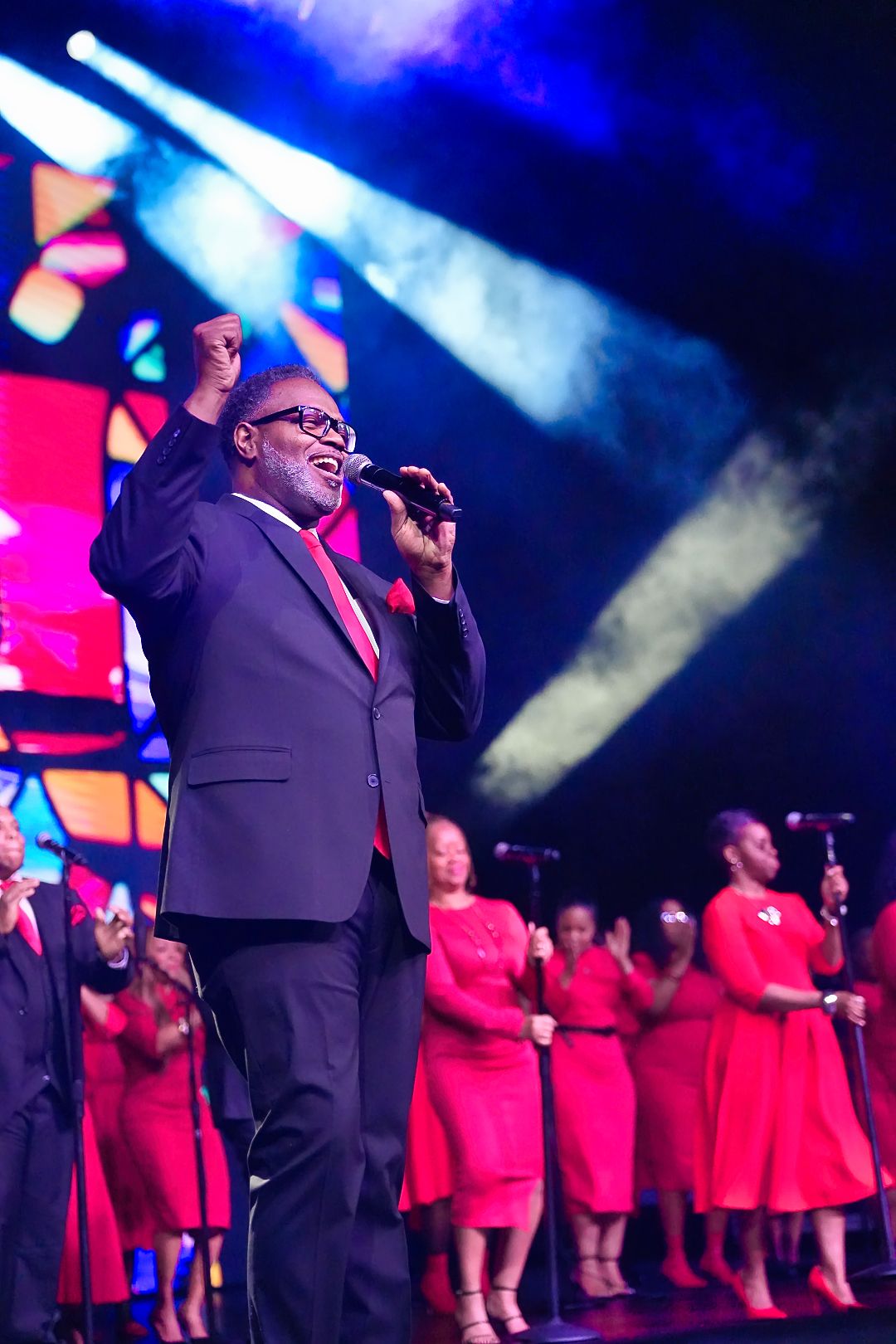 A gospel choir performer in a suit leads worship with raised hand on an illuminated stage.