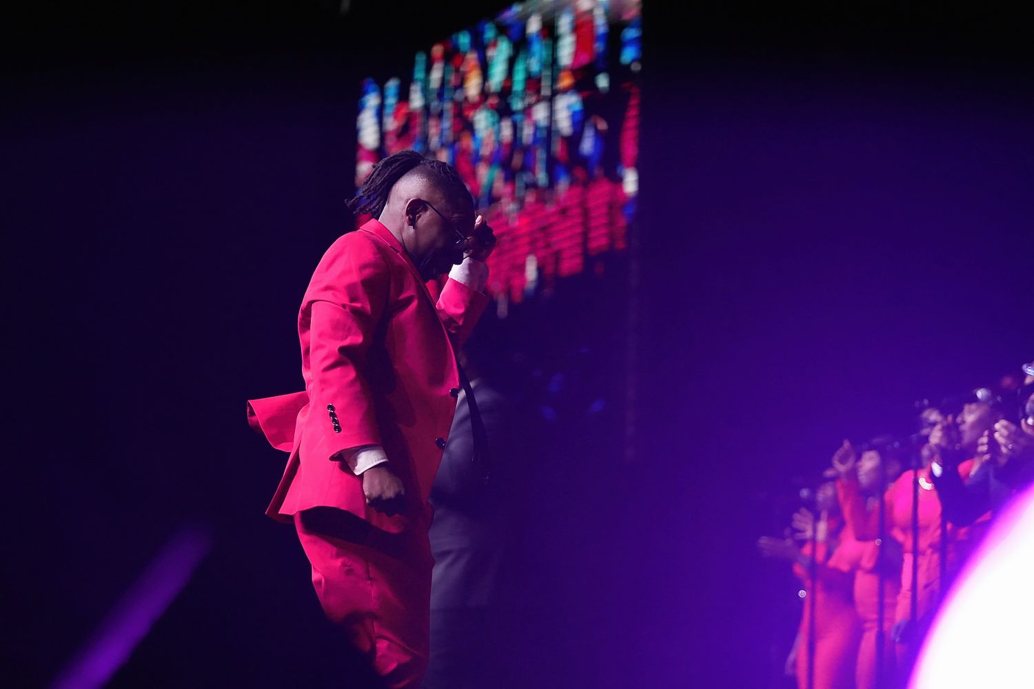 A performer in a red suit sings on stage with dramatic purple lighting.