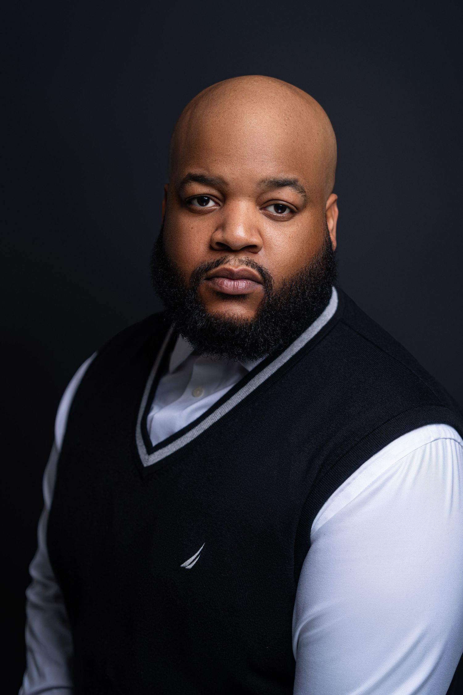 Professional studio portrait of person in black sweater vest and white dress shirt against dark background.