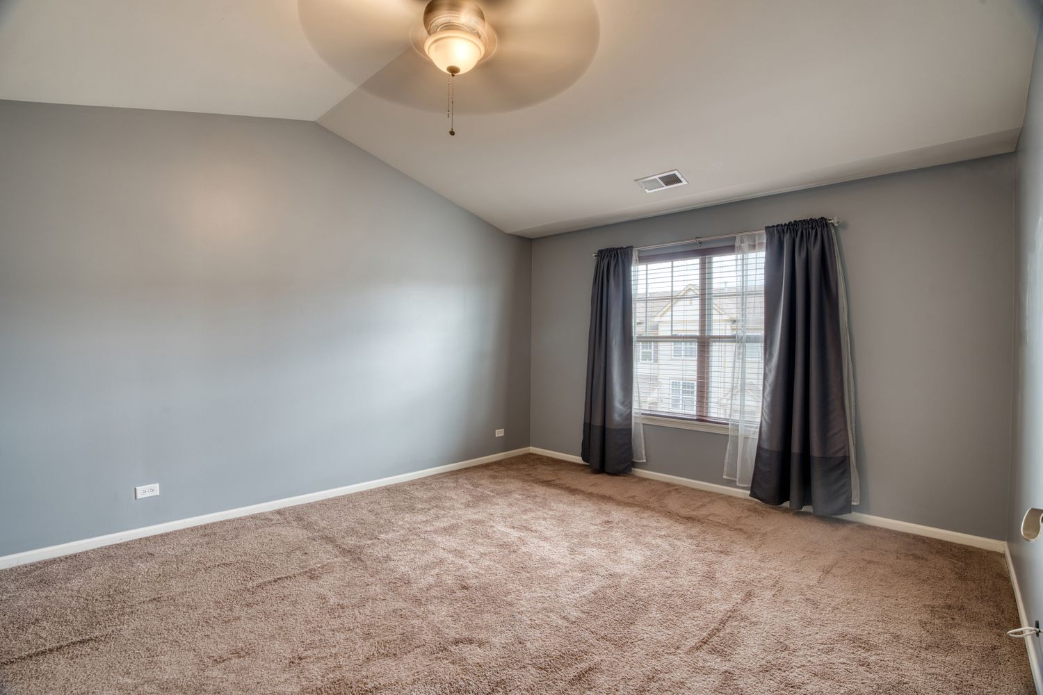 Large bedroom with grey walls, carpeted flooring, and dark curtains on windows.