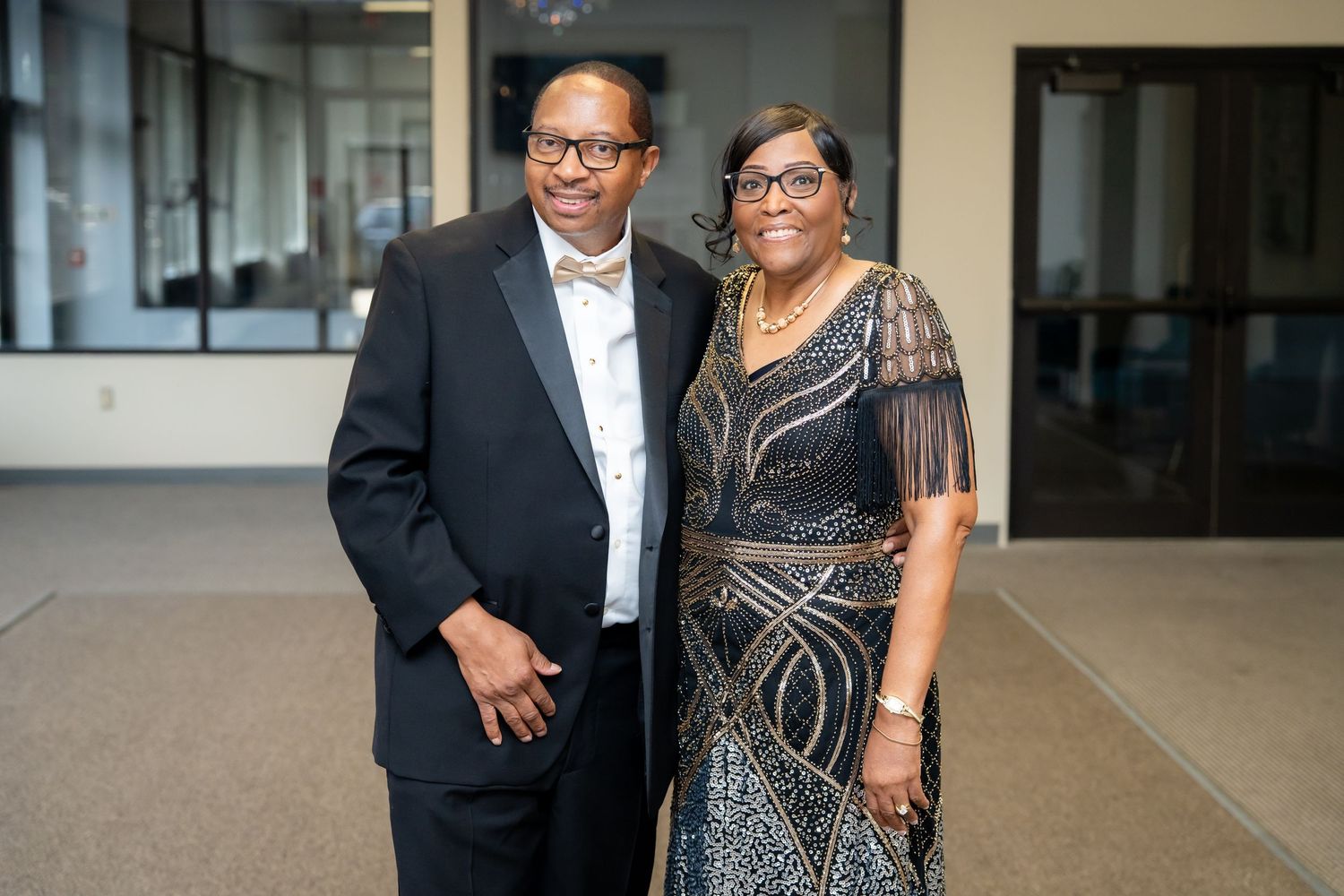 Two people in formal attire pose together at an event, with the person on the right wearing a black and gold dress.