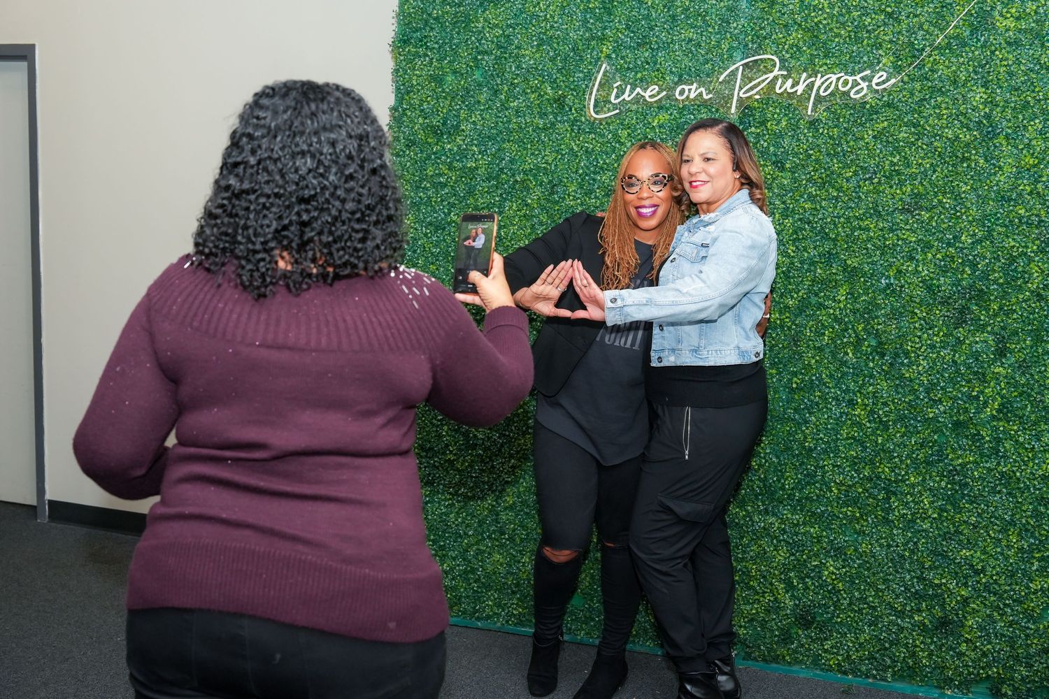 Group of attendees taking photos and interacting in front of a green foliage wall at an event.