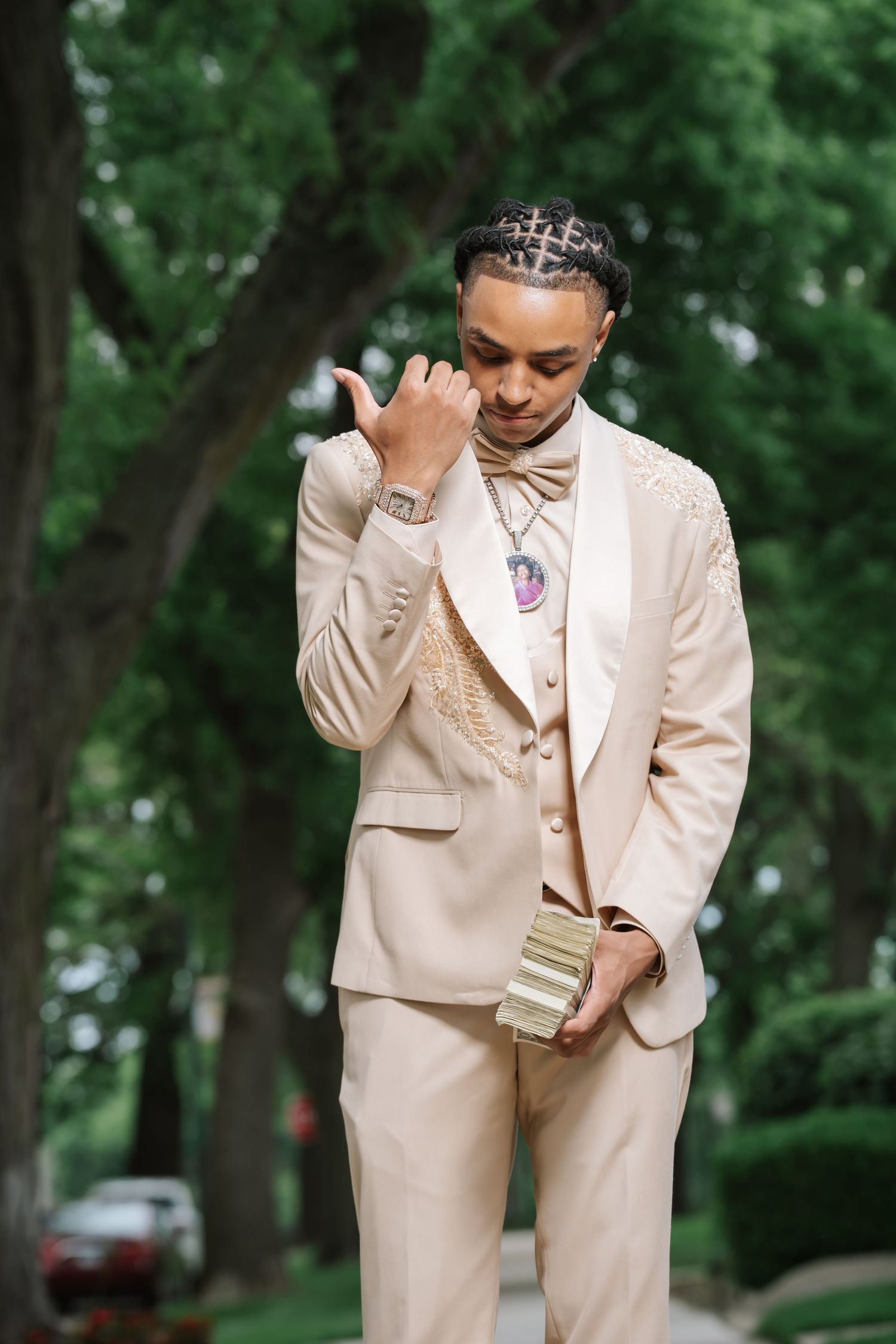 Young man in beige tuxedo suit with bow tie and gold accessories posing outdoors among green trees.