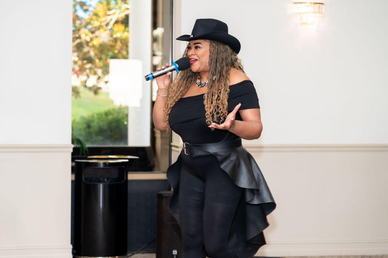 A performer in a black dress and hat sings into a microphone next to a piano during an outdoor performance.
