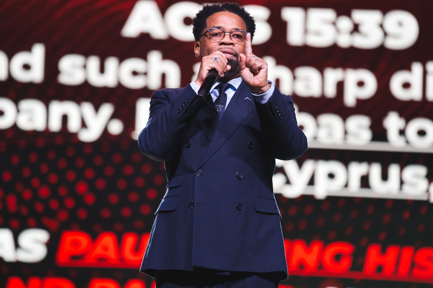 Speaker on stage with dark suit against red digital background display.