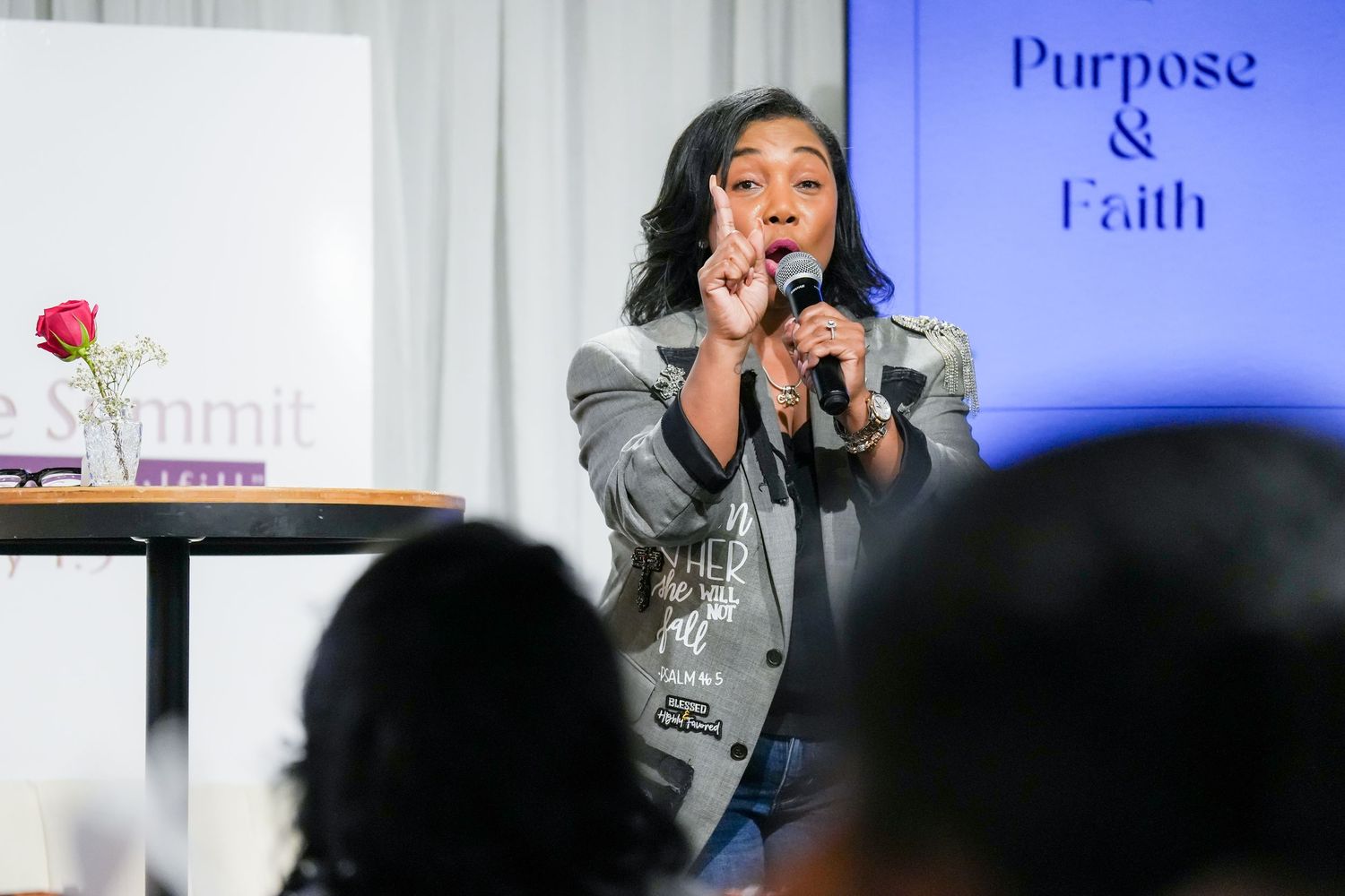 A speaker gestures while presenting on stage at a conference with blue lighting and Purpose & Faith signage visible.