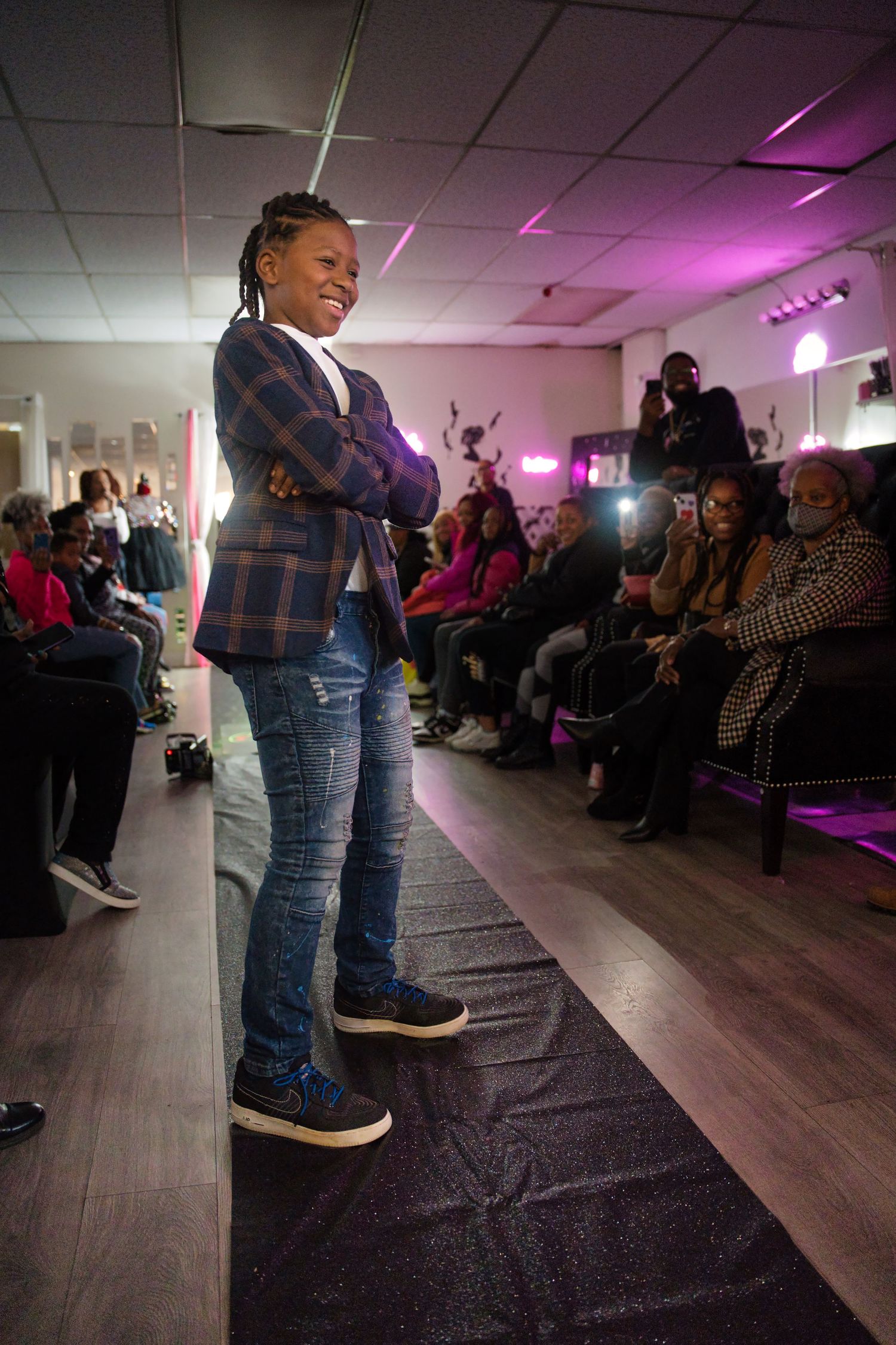 A fashion show runway with purple lighting showcases a model walking on a dark floor as audience members watch from the sides.