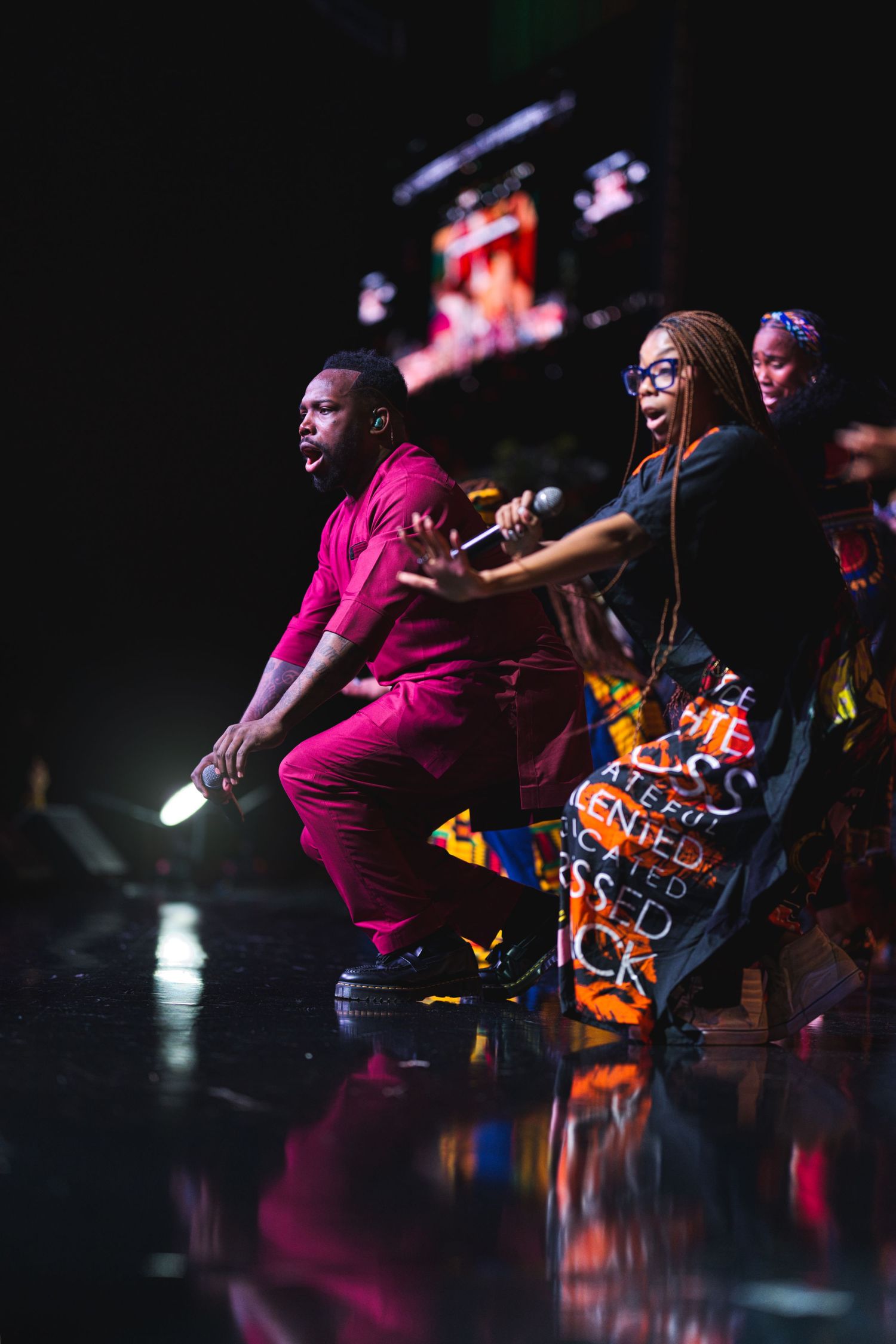 Performers on a dark stage with colorful lighting and reflective floor during an energetic show.