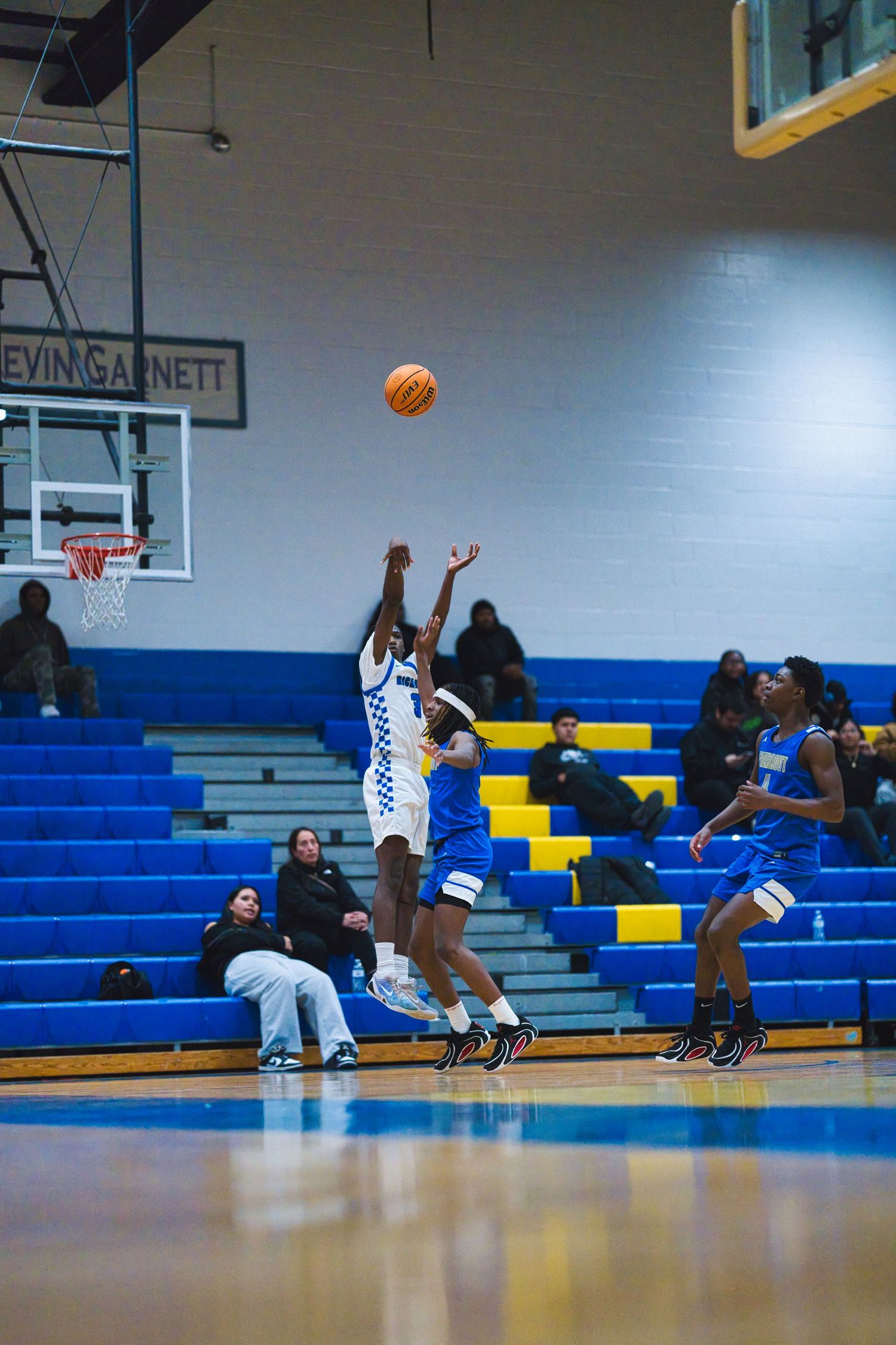 Basketball player takes a jump shot during practice in an empty gymnasium with blue and yellow bleachers.