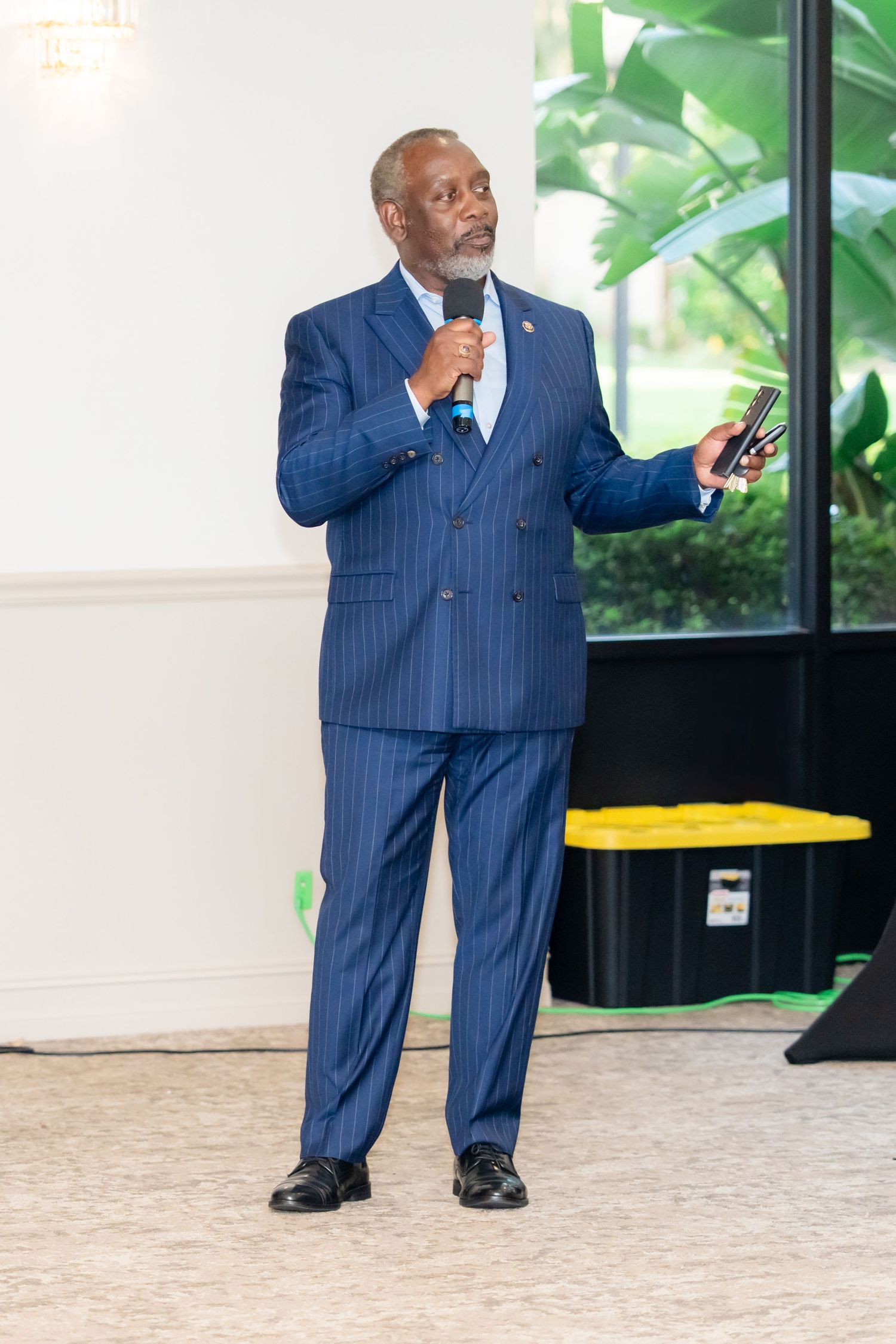 A person in a pinstriped navy blue suit stands in a room with green foliage visible in the background.