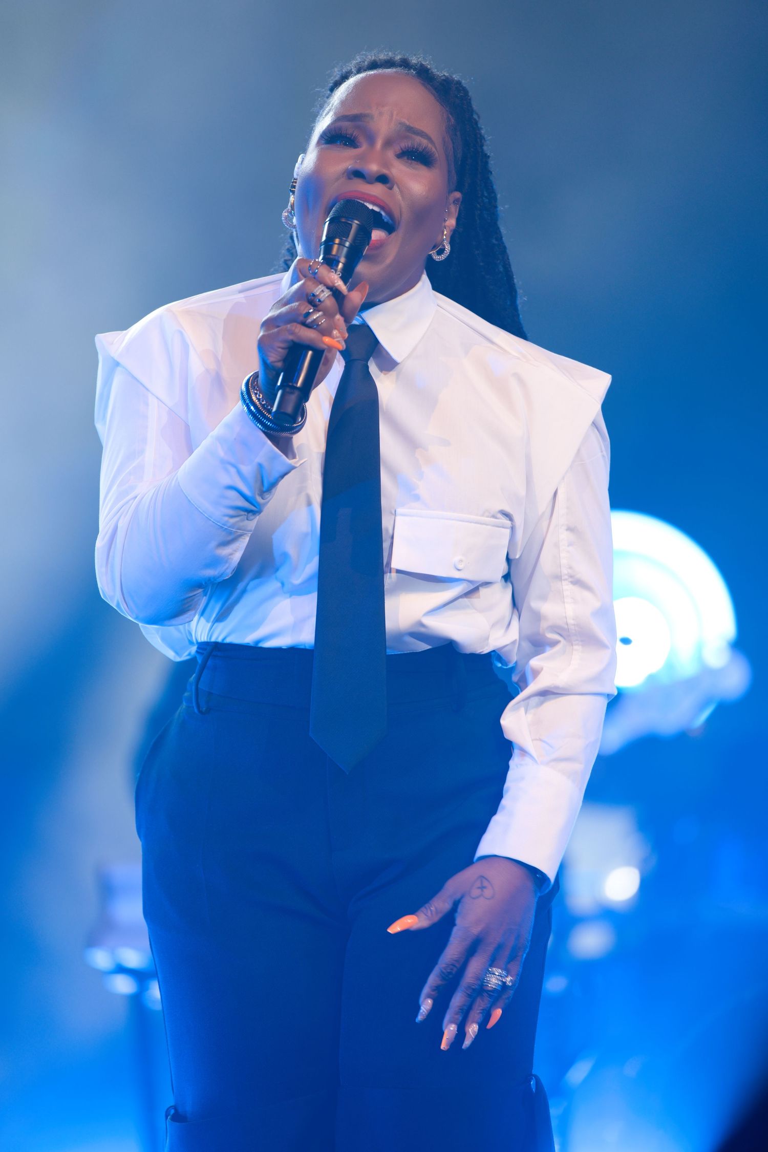 A performer in a white jacket sings passionately on stage with dramatic blue lighting in the background.