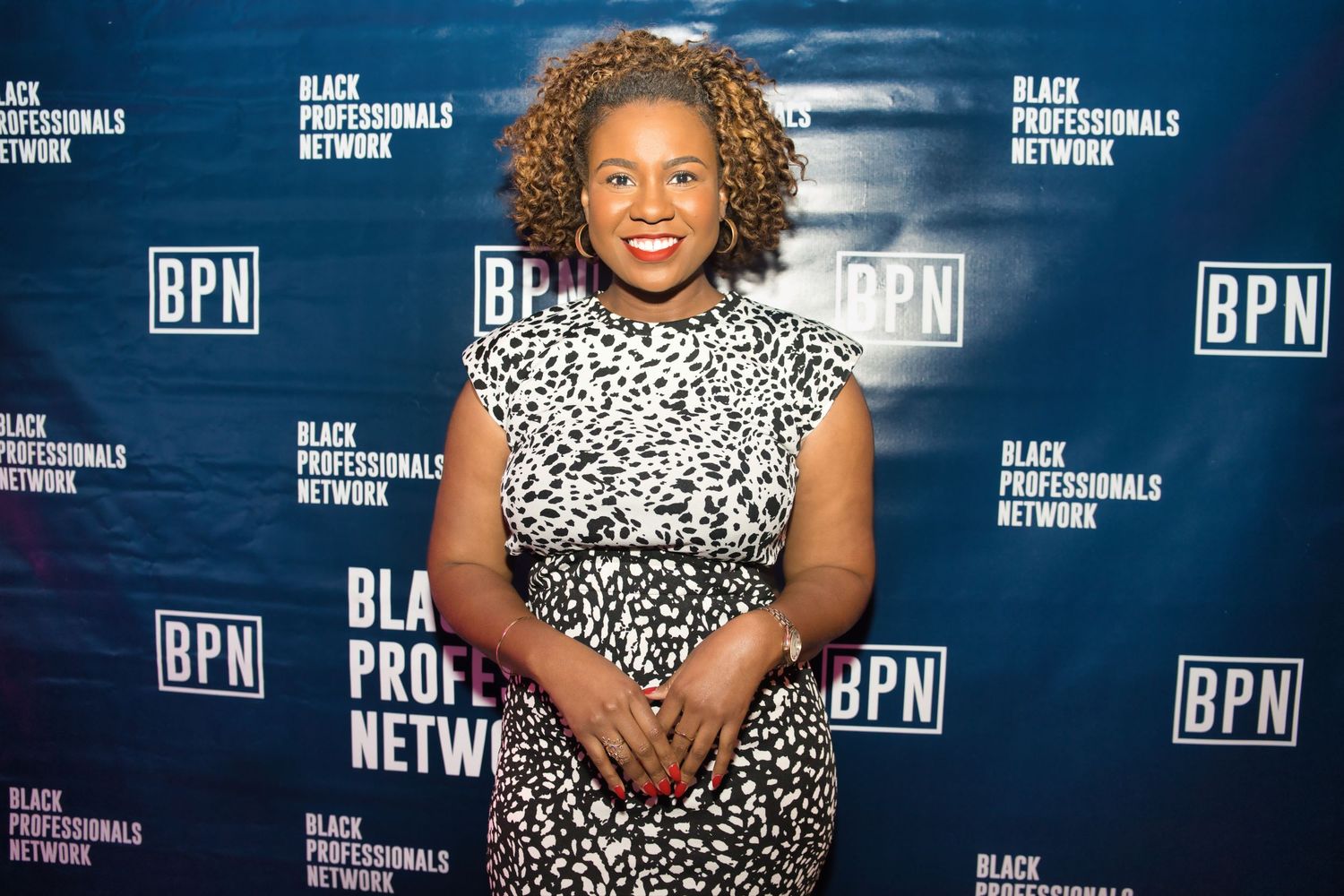 A person in a black and white patterned dress poses in front of a BPN step and repeat media wall.