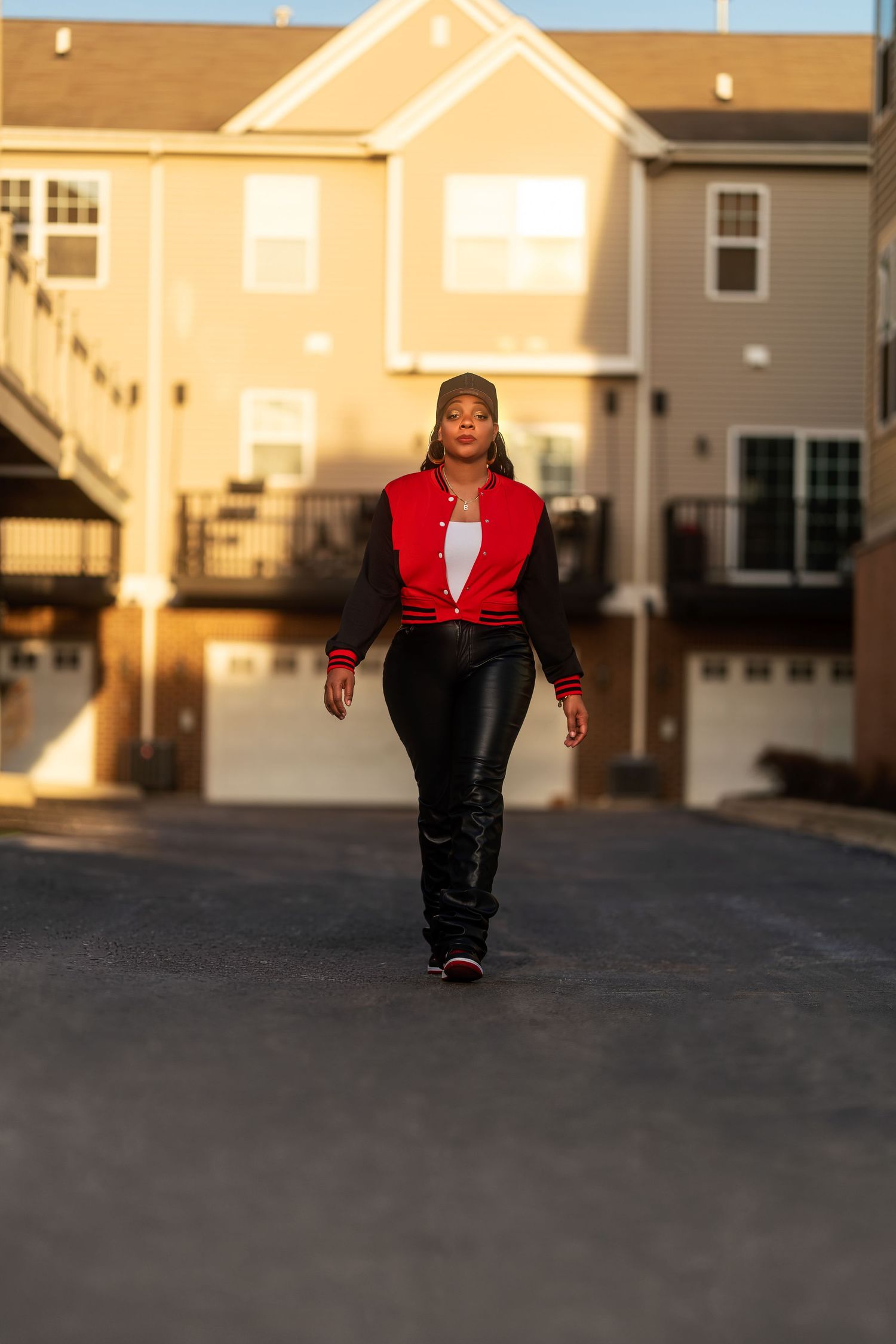 Woman in red varsity jacket and black leather pants walking confidently down a residential driveway at golden hour.