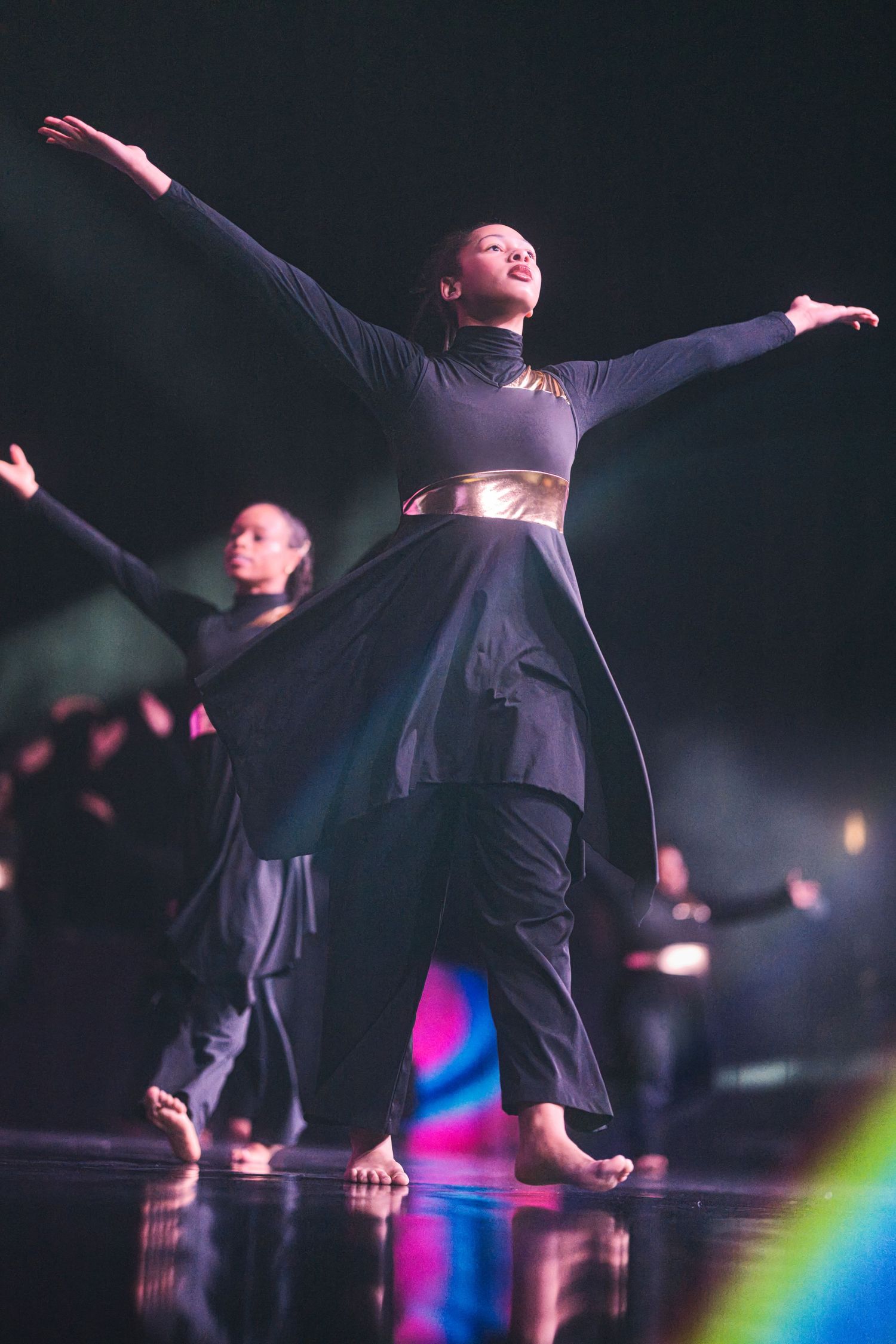 Performers on stage dance with outstretched arms in dramatic lighting with colorful rainbow effects in the background.