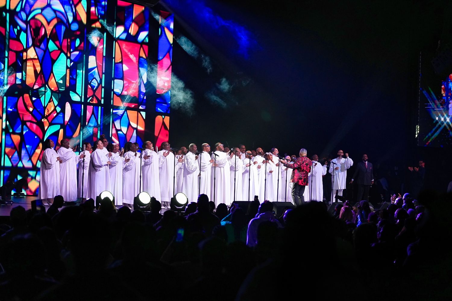 A choir in white robes performs on stage with dramatic lighting and stained glass backdrop.