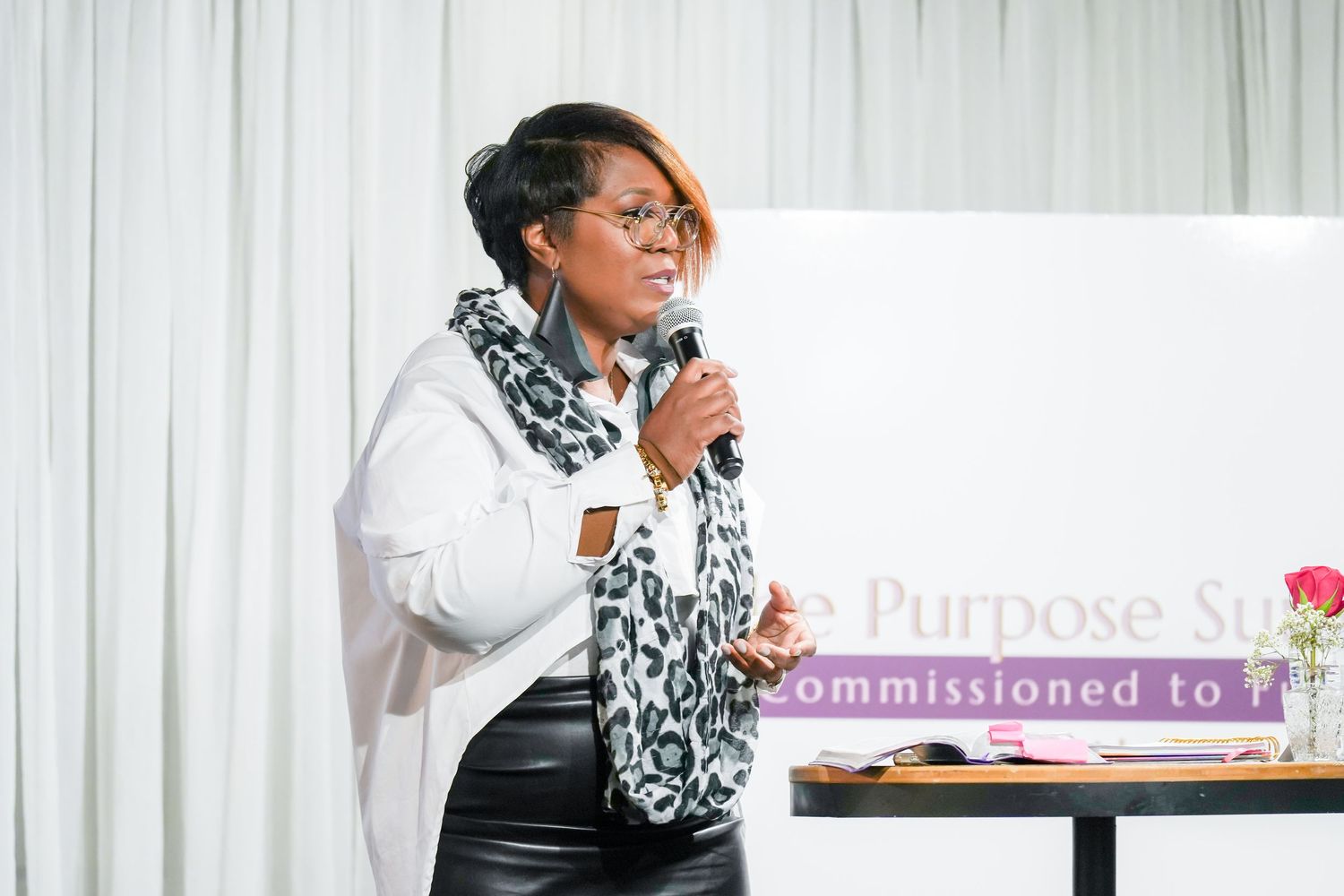 Professional speaking at a business event with white curtain backdrop and presentation materials.