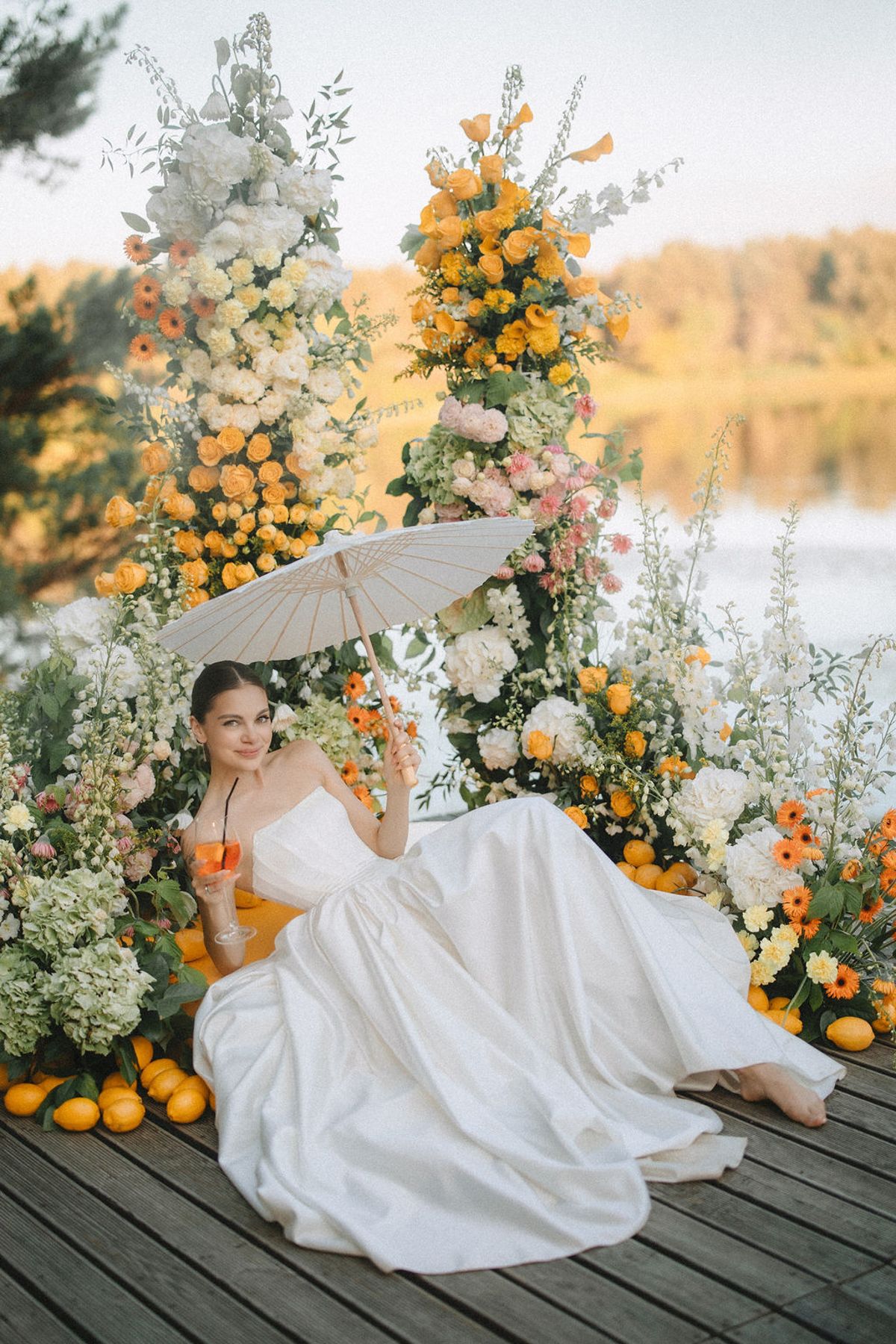 Romantic bridal photoshoot on wooden deck with orange citrus fruits and white floral arrangements creating dreamy atmosphere.
