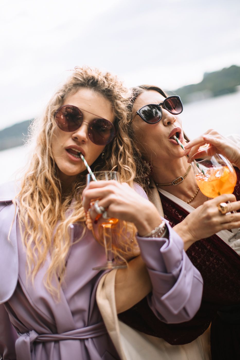 Two friends in sunglasses enjoying orange cocktails outdoors on a sunny day while wearing casual attire.