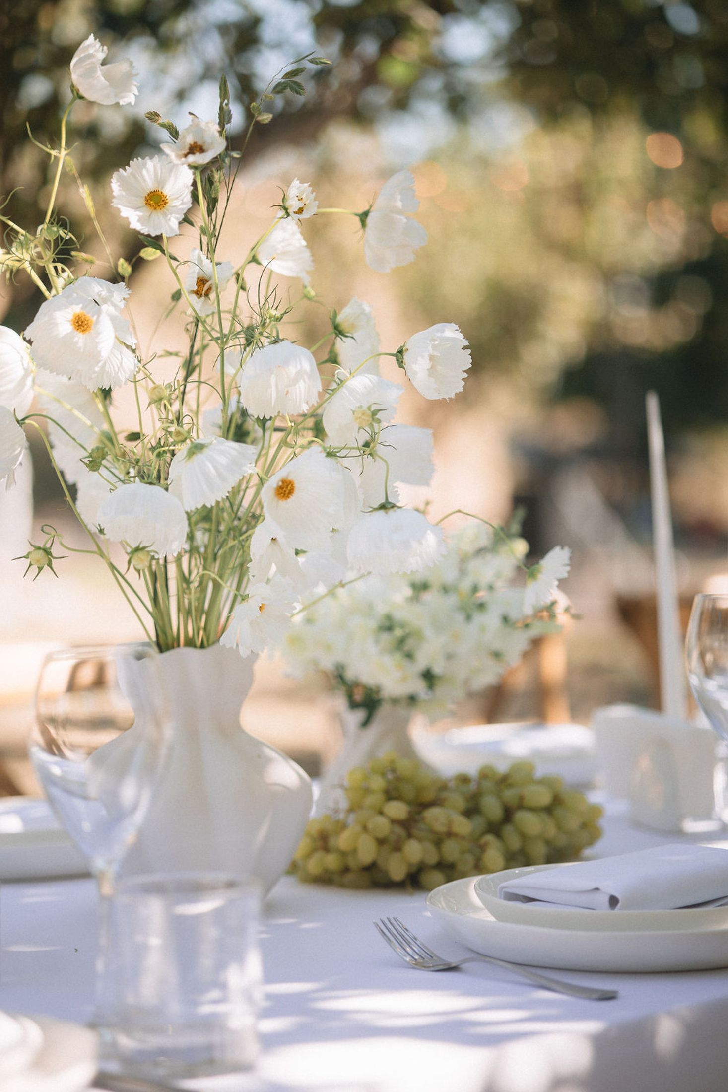 White floral centerpieces with delicate blooms arranged in elegant teacups on a sunlit wedding reception table.