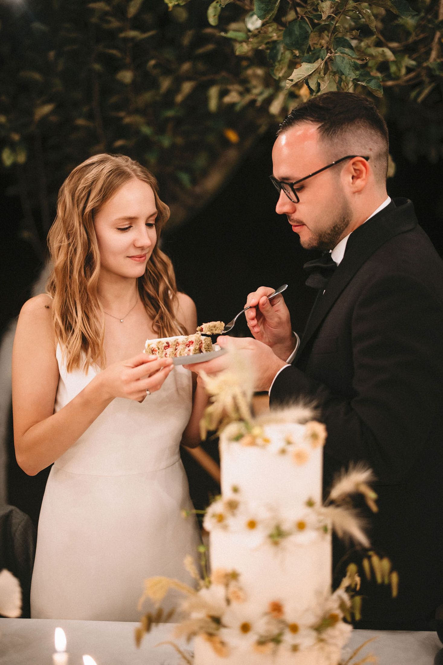 Newlywed couple sharing bites of wedding cake in a romantic outdoor setting with natural lighting.