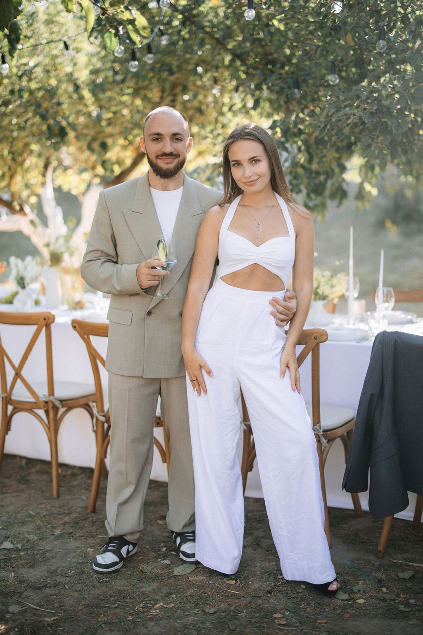 Couple in white and beige attire pose together at an outdoor wedding reception with rustic wooden chairs.