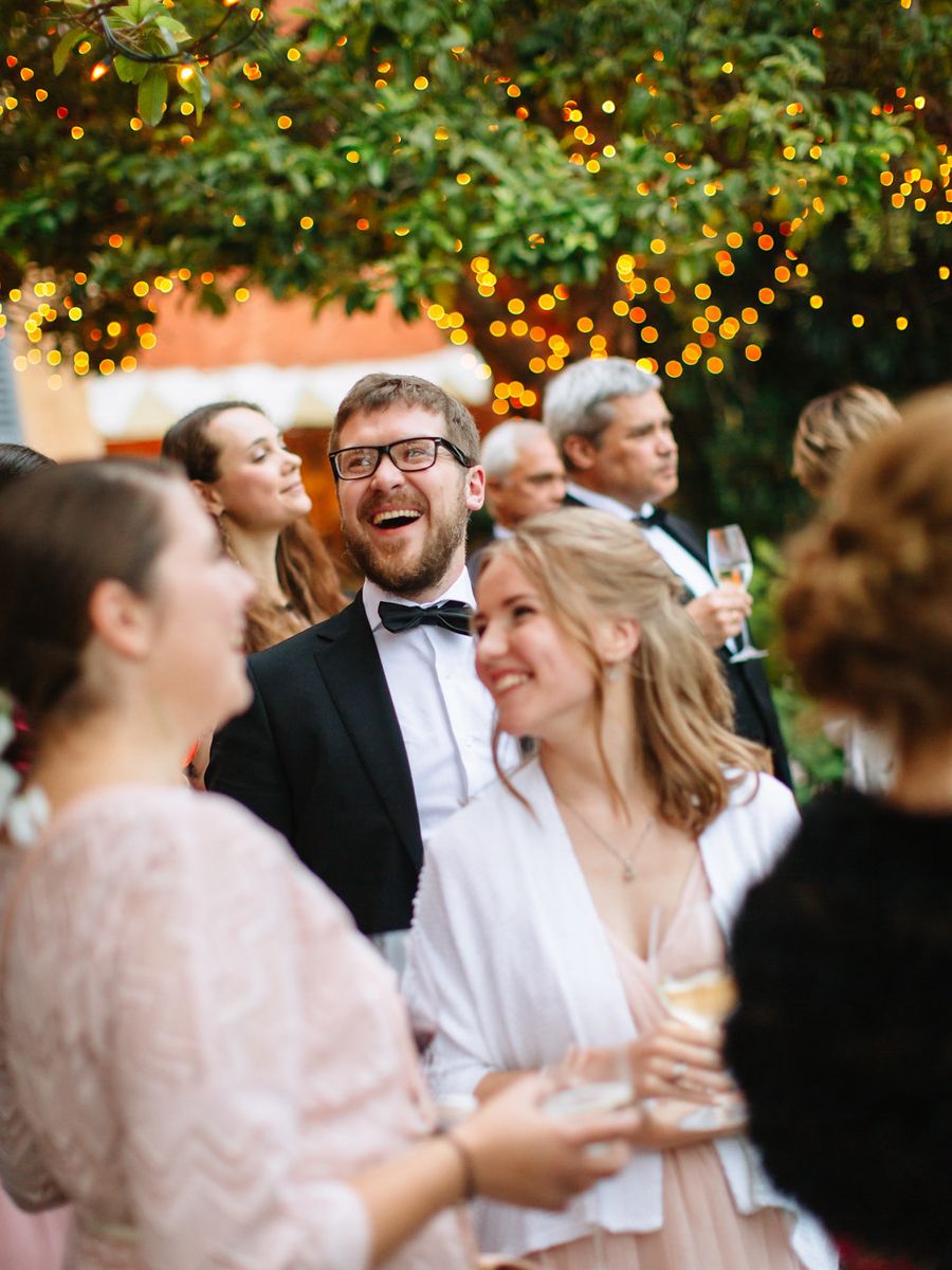 Wedding celebration with guests mingling under trees decorated with warm glowing string lights at dusk.