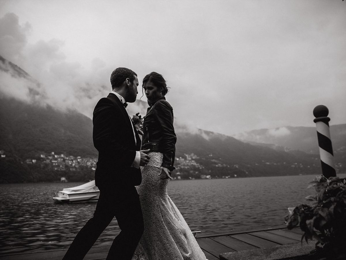 A romantic couple embraces on a misty lakeside dock with mountains in the background in black and white.