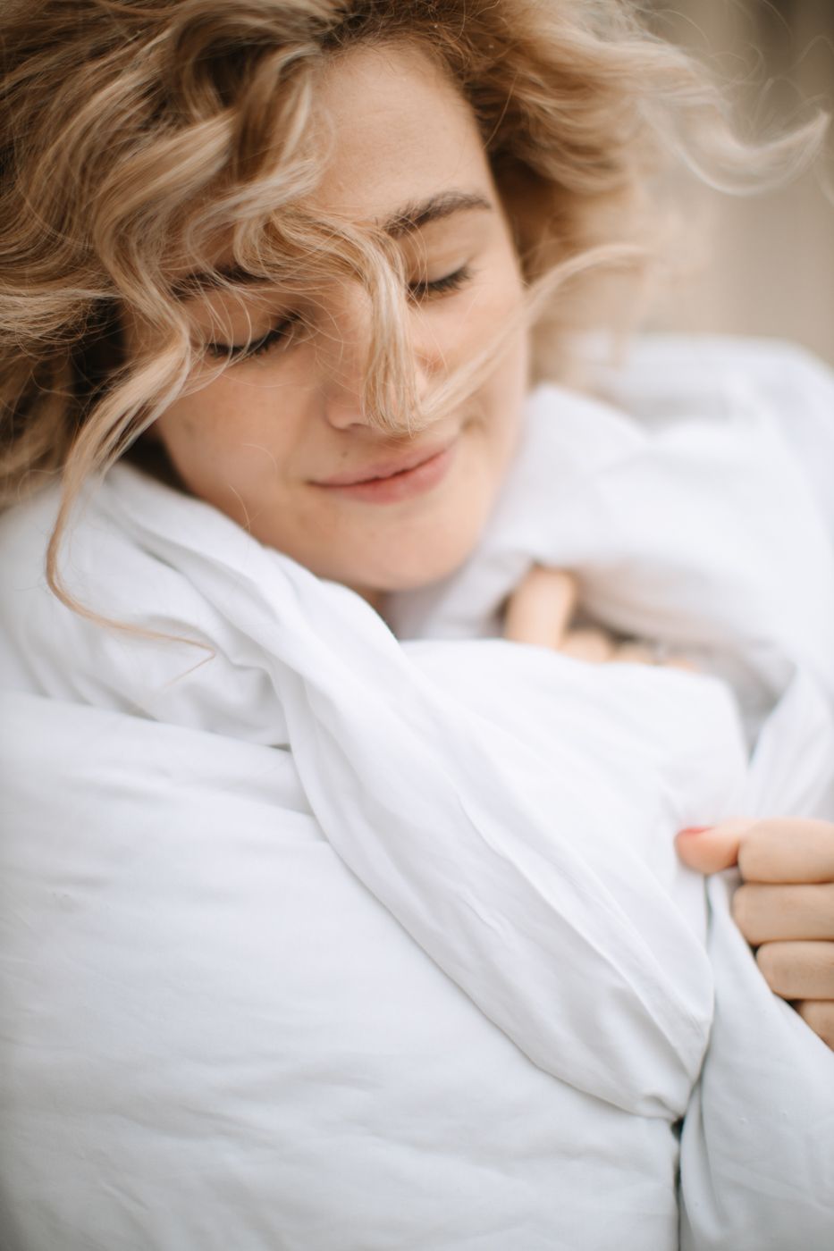 Serene portrait in soft lighting showing person wrapped in white bedding with gentle expression.