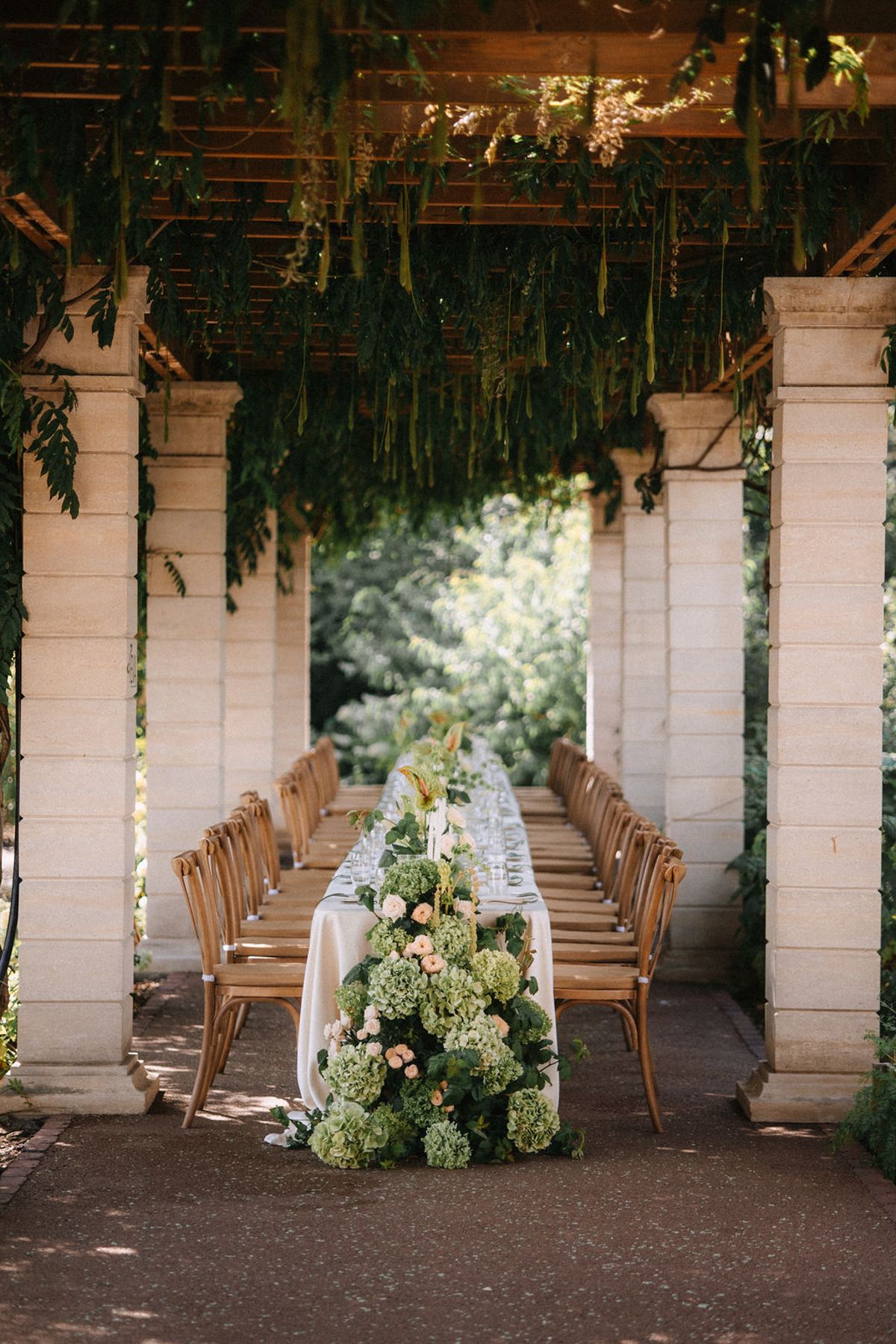White wooden pillars and chairs create an elegant outdoor wedding ceremony space with hanging greenery and floral arrangements.