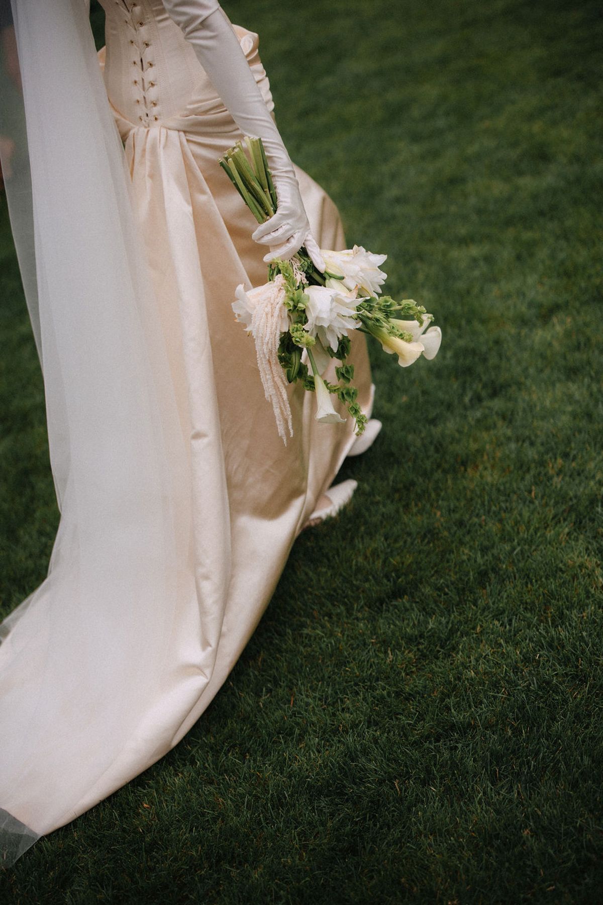 A flowing white wedding dress hem and delicate lace details trail gracefully across green grass in an artistic composition.