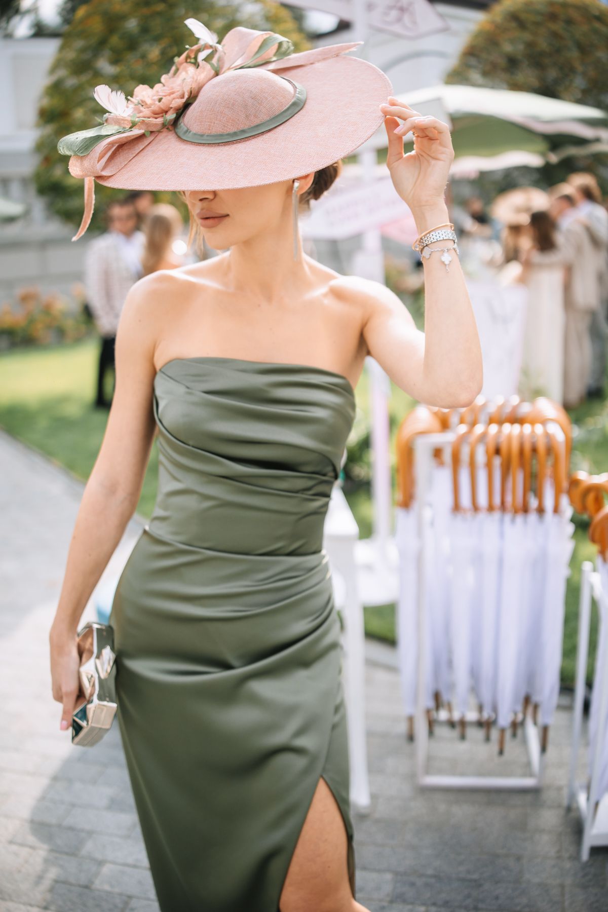 A person in an olive green dress and pink hat dances elegantly at an outdoor event with white chairs in the background.