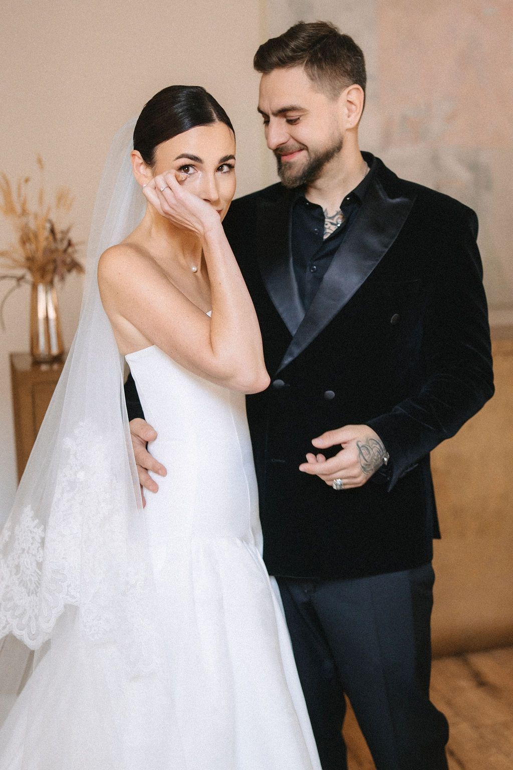 A couple shares an emotional moment on their wedding day, with the bride in white and groom in a black velvet jacket.