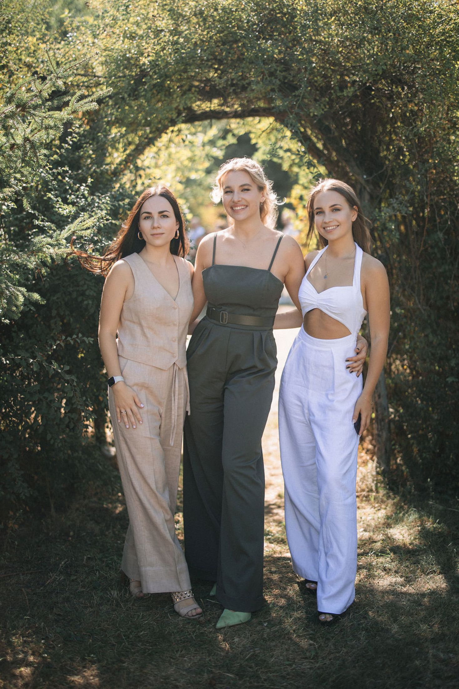 Three people wearing casual summer outfits pose together outdoors against green foliage in natural lighting.