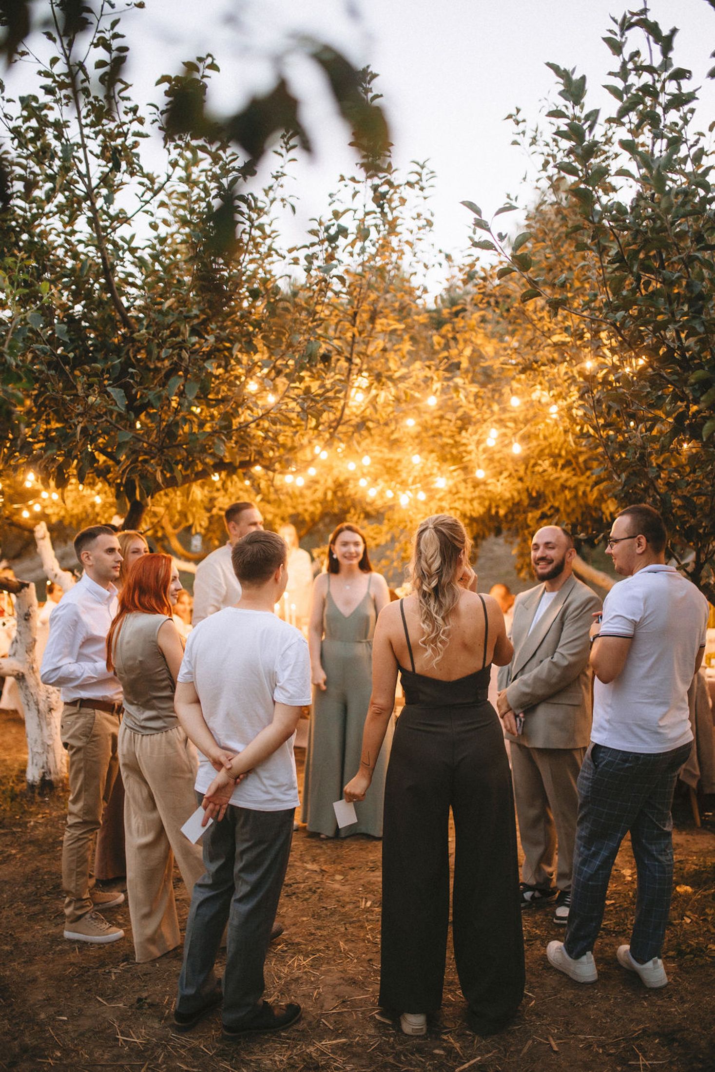 A group of people gather outdoors at sunset with string lights glowing through autumn trees in the background.