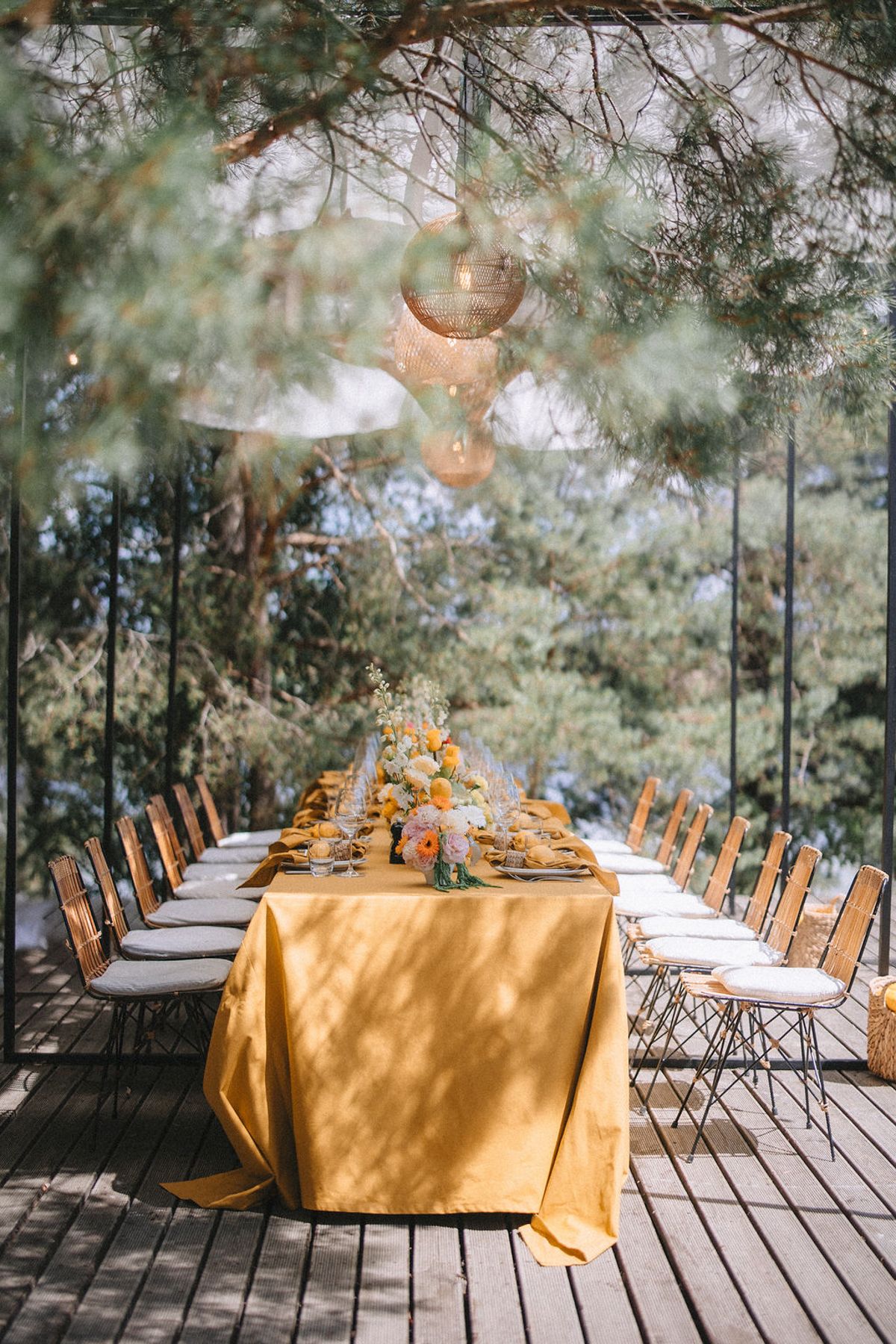 Elegant outdoor dining setup with gold tablecloths and wooden chairs under hanging lanterns and tree canopy.