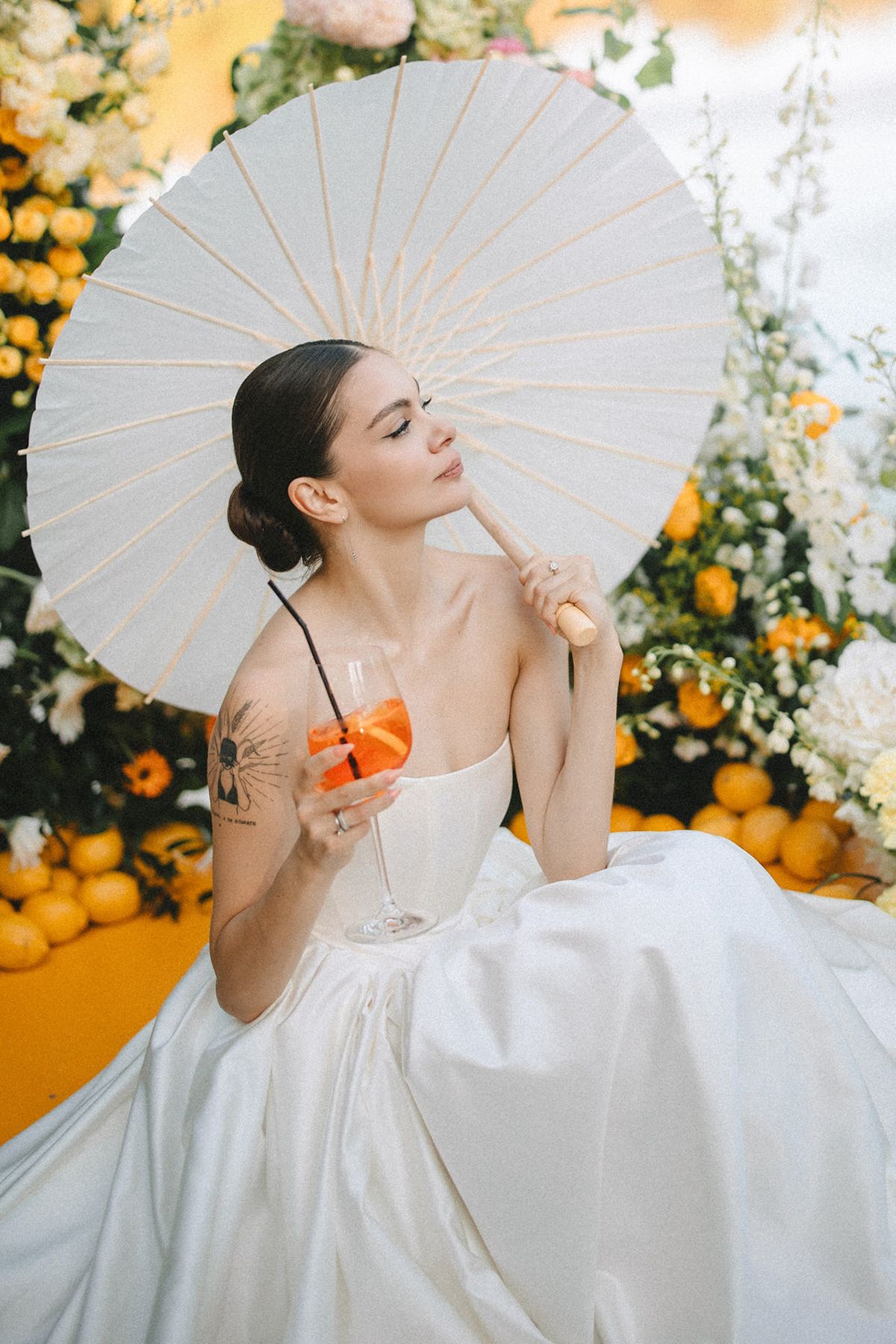 Elegant figure in white wedding gown poses with paper parasol and orange flowers in garden setting.
