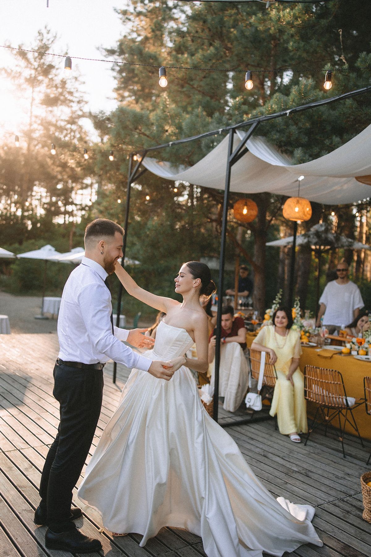 A joyful couple dances together during their outdoor wedding reception under string lights and white canopies at sunset.